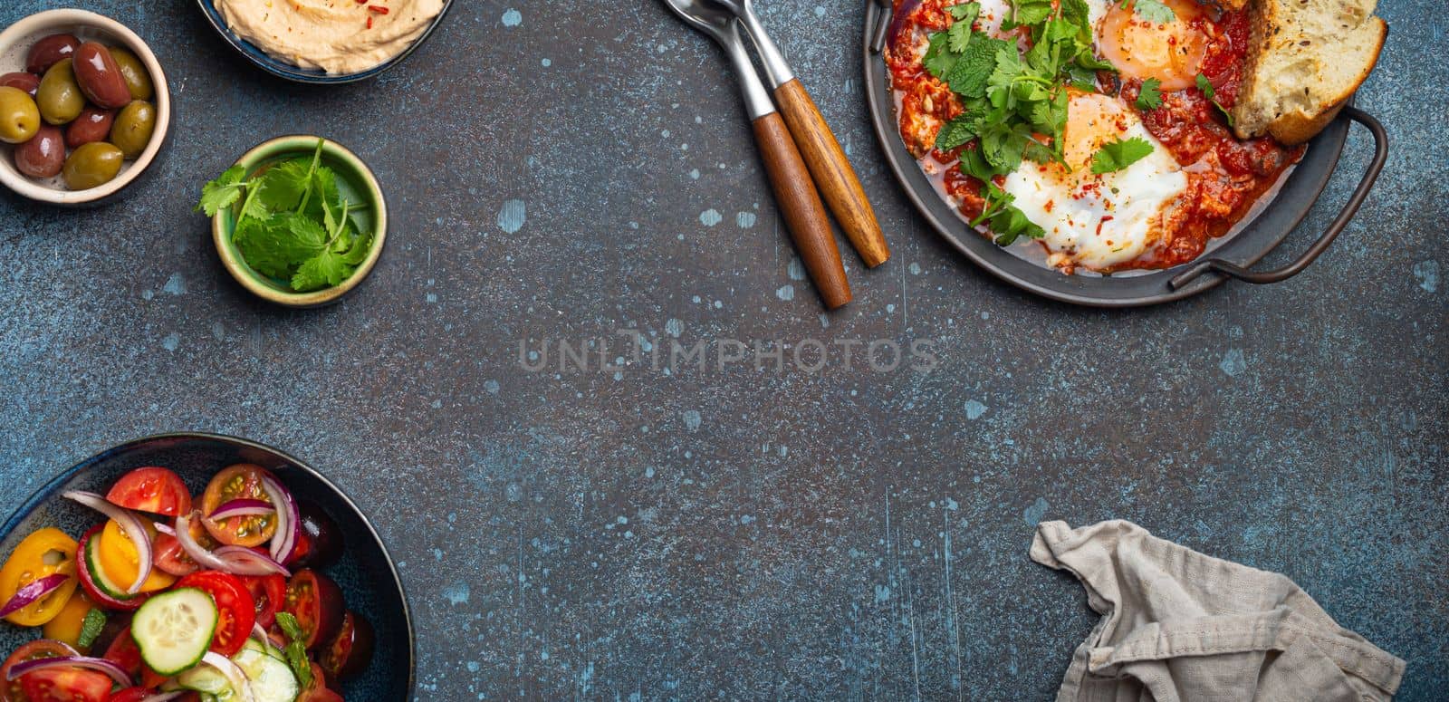 Middle Eastern traditional breakfast or brunch with eggs Shakshouka in pan with toasts, fresh vegetables salad, hummus and olives on rustic concrete background table from above, space for text