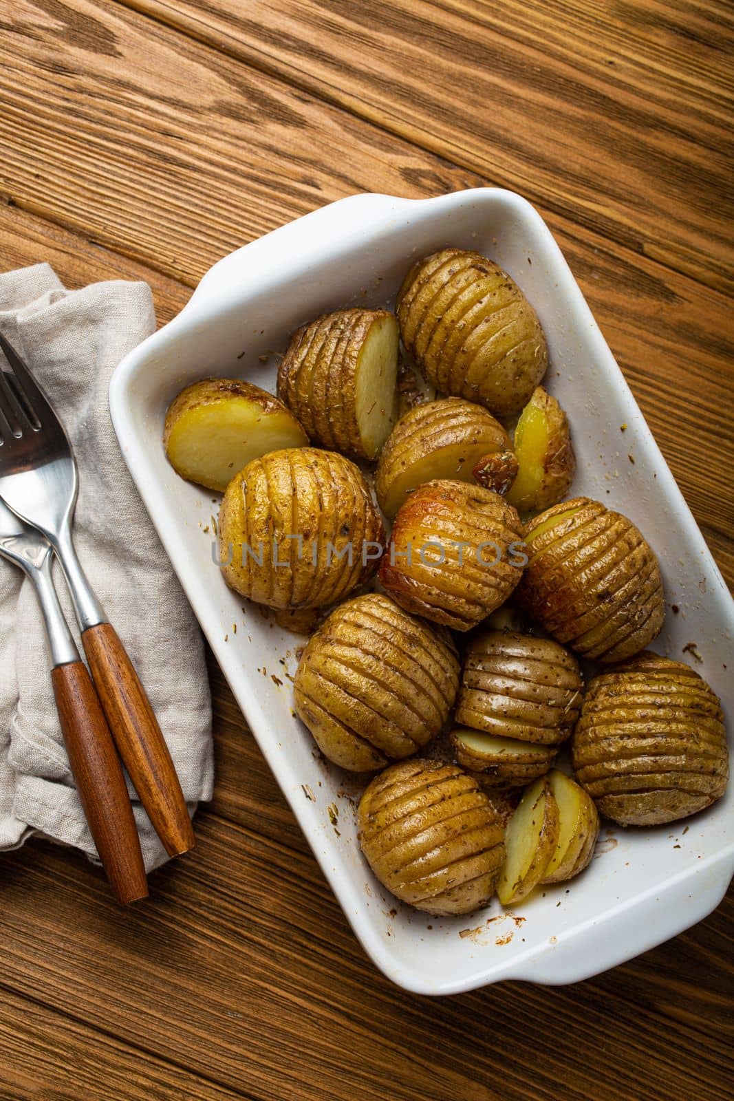 Hasselback potatoes with herbs in white ceramic casserole dish on wooden rustic table background from above. Delicious homemade accordion baked potatoes for dinner
