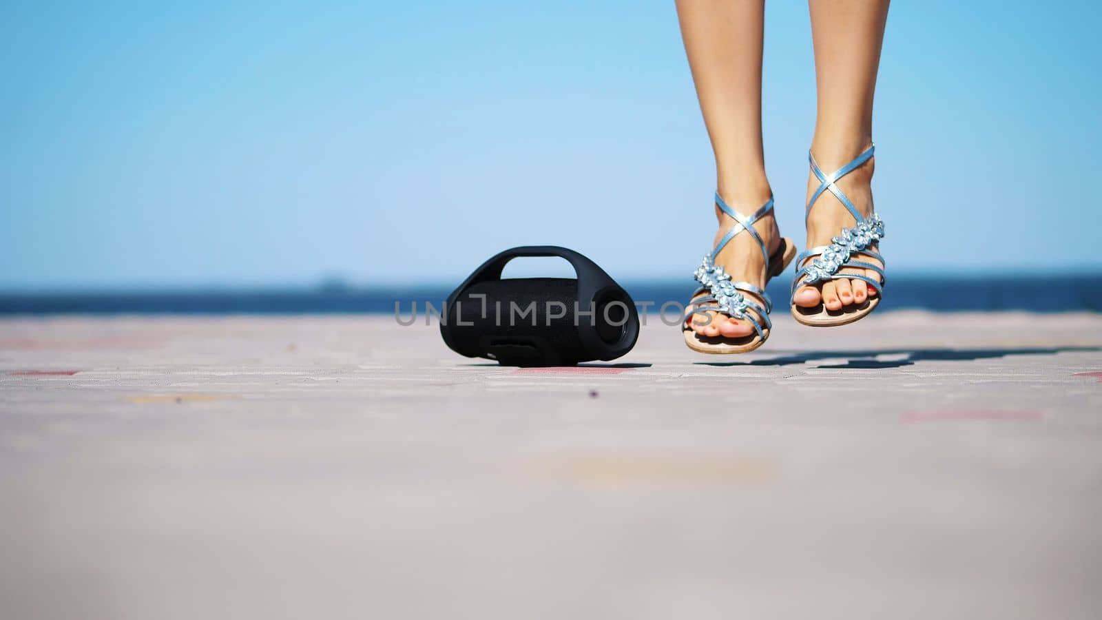 close-up, mini music bluetooth portable black cylinder wireless loudspeaker. female legs in silver sandals, with bright red pedicure, dancing to the music. on the beach, in summer. High quality photo