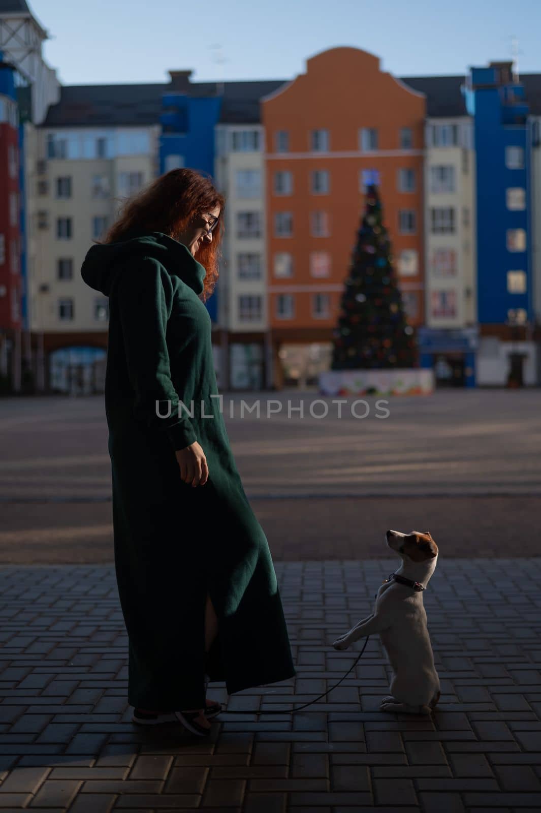 Red-haired woman in a green dress with a dog Jack Russell Terrier in the square against the background of the Christmas tree