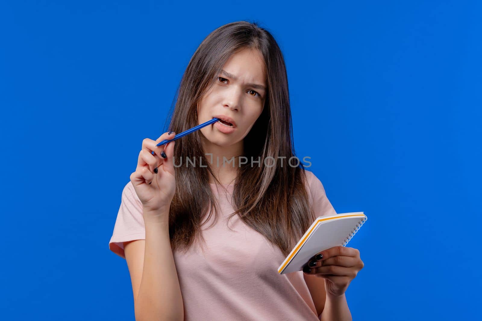 Young woman writing notes in planner with pen. Student girl thinking about future plans and to-do list in notebook for week, month, year. Keeping personal diary on blue background. by kristina_kokhanova