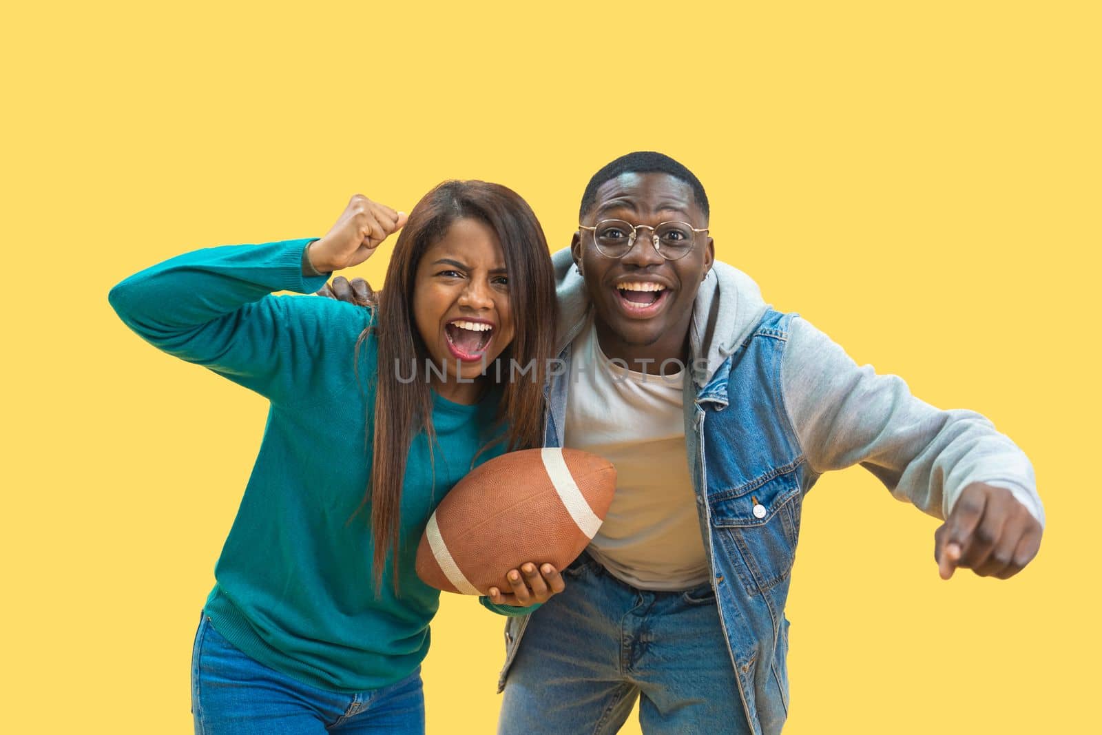 Excited ethnic diversity couple enjoying sports together isolated on a yellow background looking at camera with a American football ball in their hands. Happy football fans concept. High quality photo