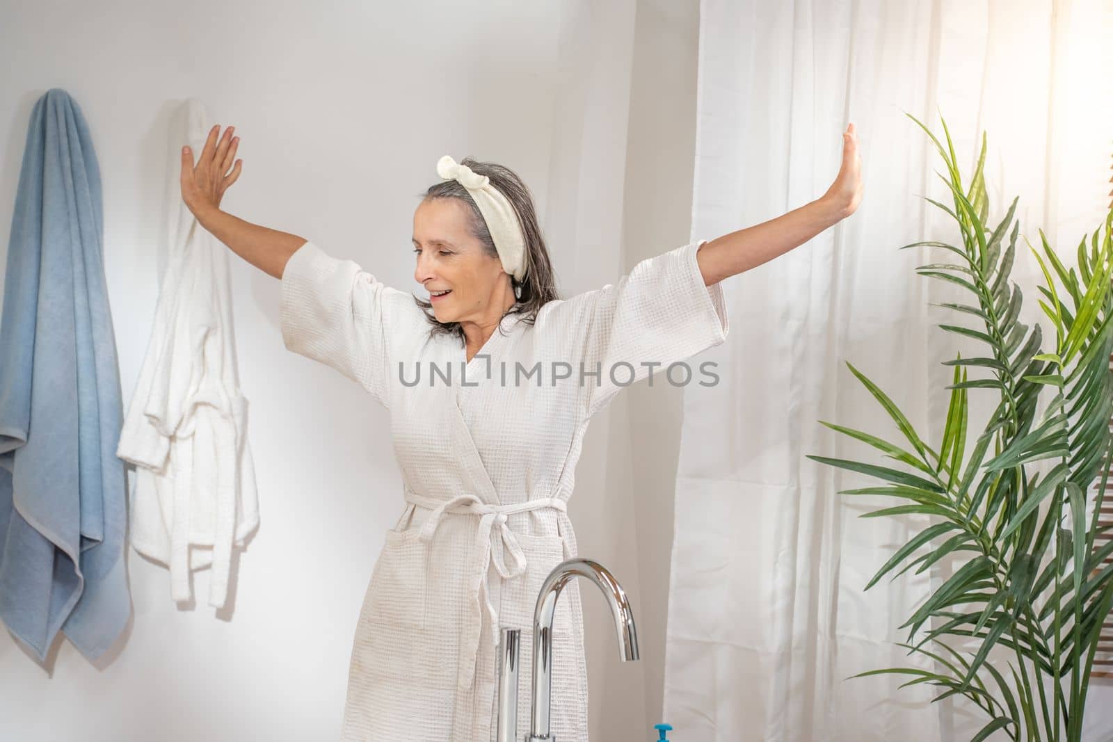 Happy smiling beautiful middle aged woman in bathrobe stretching out arms at luxury bathroom. Spa procedures anti age recreation skin and body care, cosmetic and beauty concept.