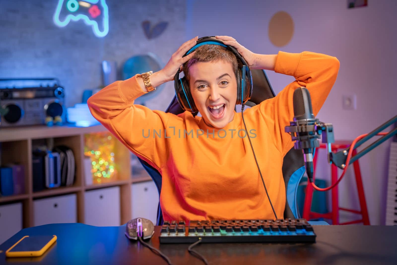 Excited Gamer Girl with hands in her head surprised using Headphones with a Mic Playing Online Video Game on Personal Computer. Colorful neon lights room. High quality photo