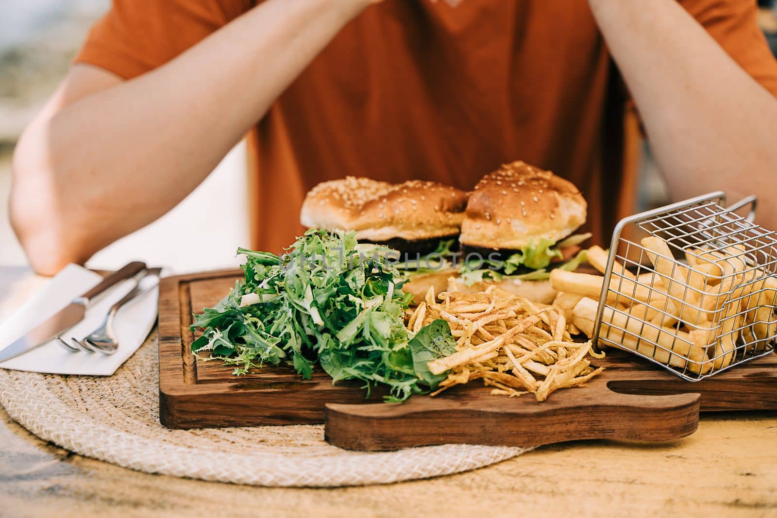 Cropped closeup man male sitting in front of sliced grilled tasty beefy cheese burger hamburger and french fries on a plate in the street cafe. Copy blank space.