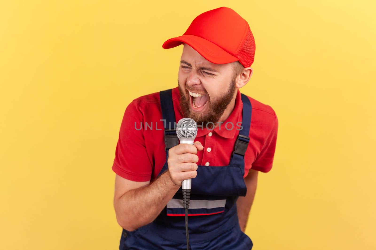 Portrait of extremely excited worker man wearing blue uniform and red cap relaxing at a corporate party singing karaoke at work. Indoor studio shot isolated on yellow background.