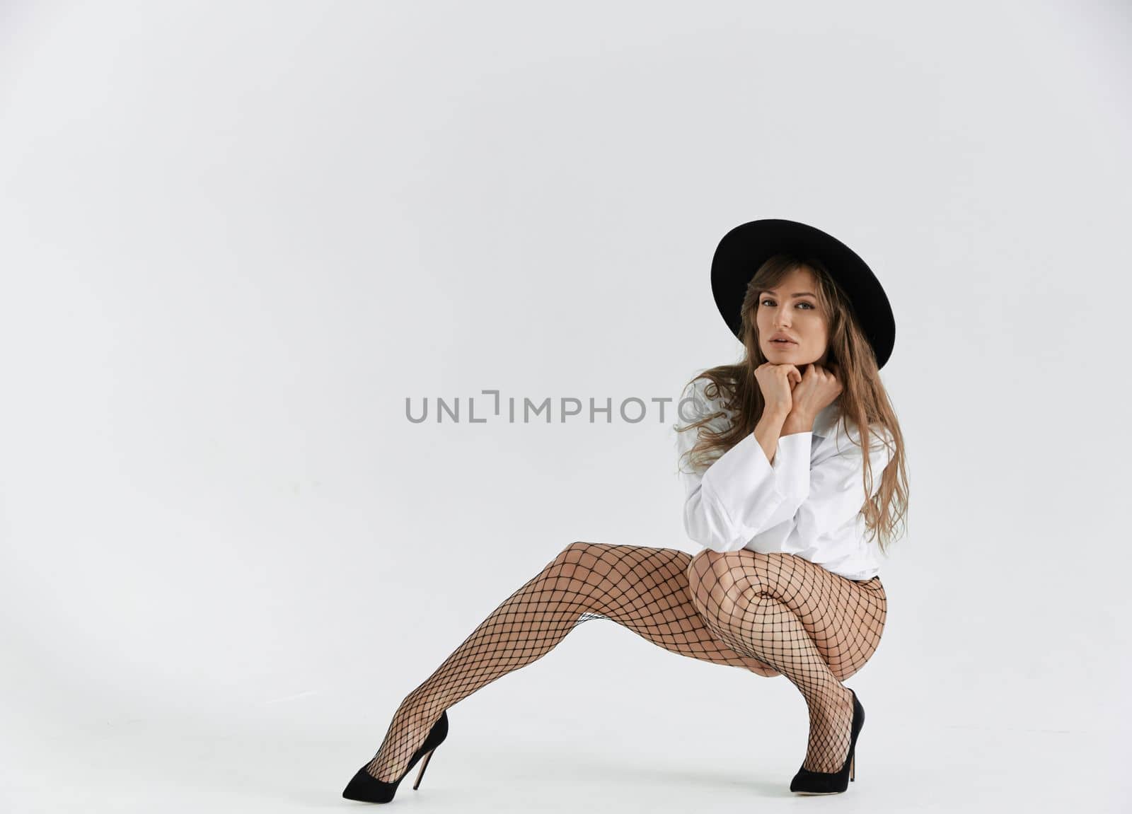 Beautiful brunette girl flirts with the camera, she is dressed in mesh tights, white shirt and black hat, long hair, pure white background, soft light by vladimirdrozdin