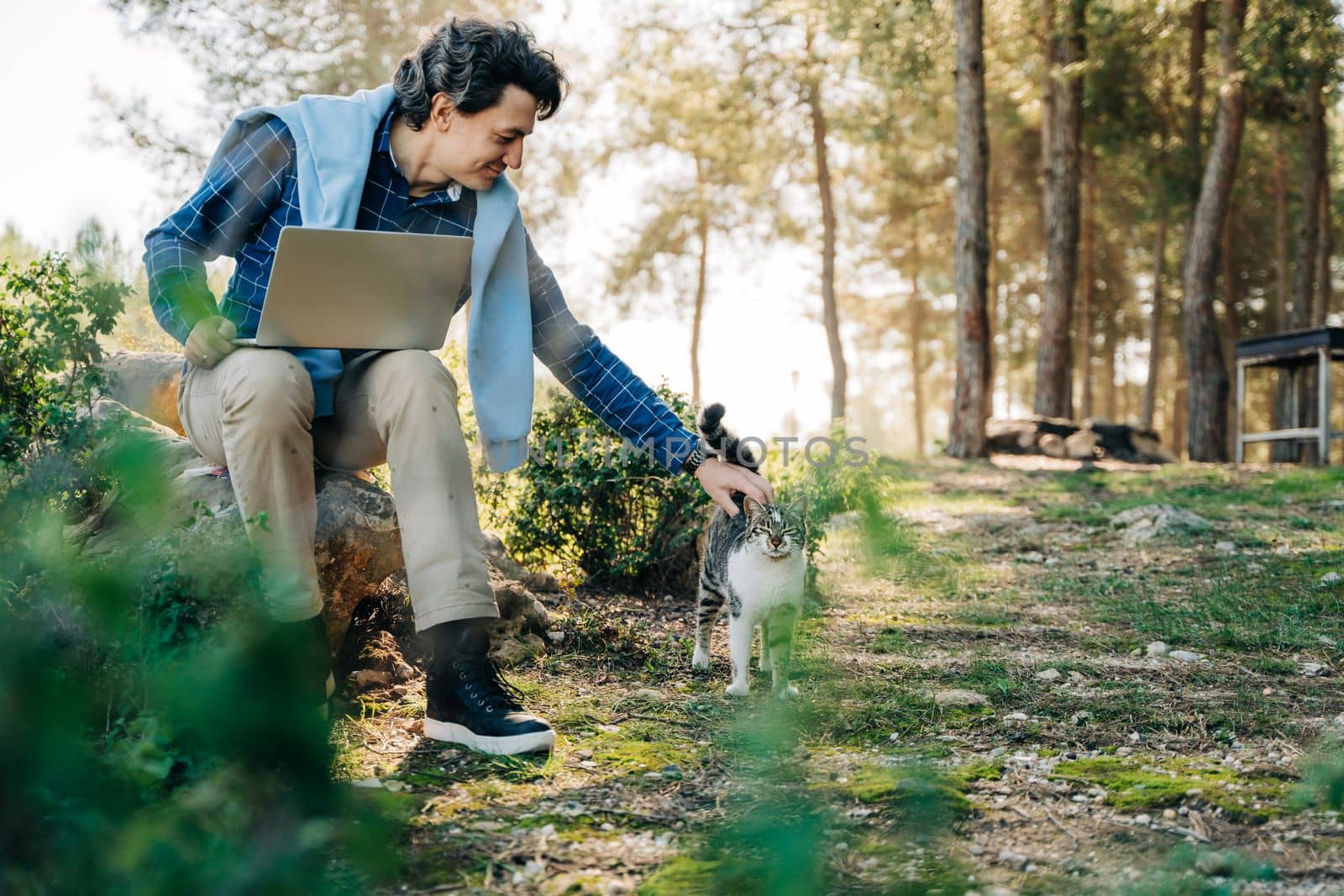 Happy cheerful man with a laptop sitting outdoors on rock in nature forest park and pet cat. Freelancer with computer typing, blogging, browsing in vacation. Freelance, distance work concept.