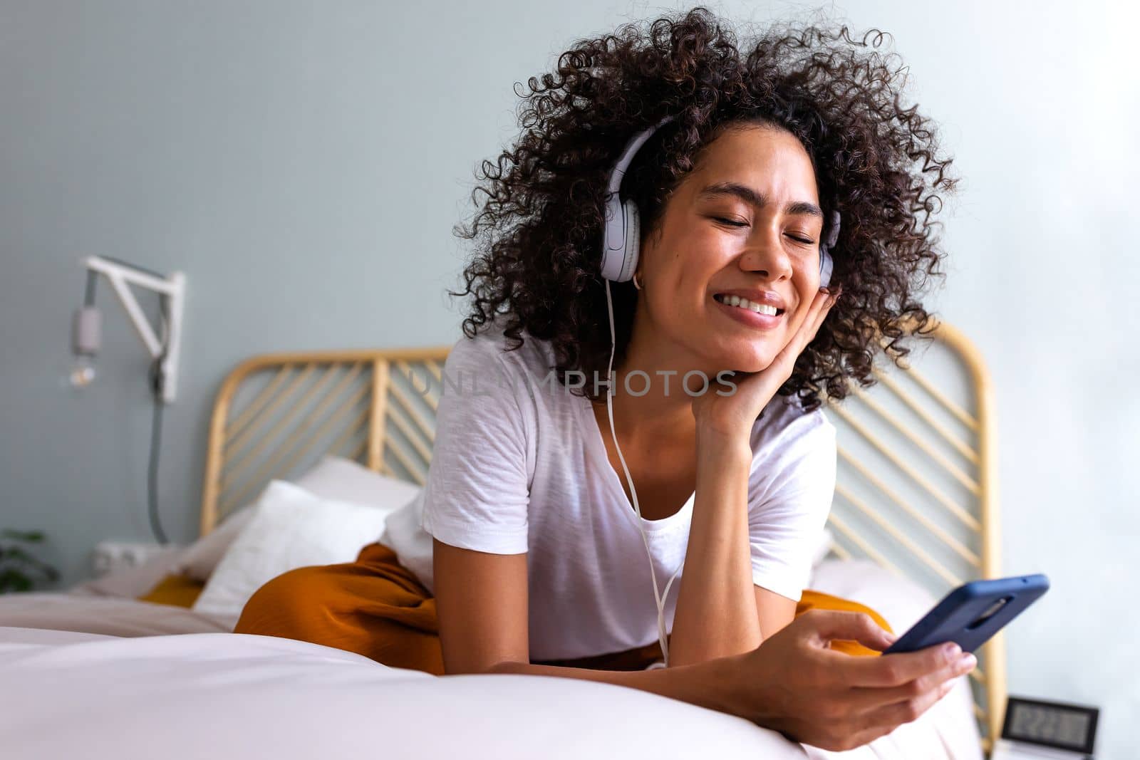 Young multiracial woman enjoying music using headphones and mobile phone relaxing on bed at home cozy bedroom. Lifestyle concept.