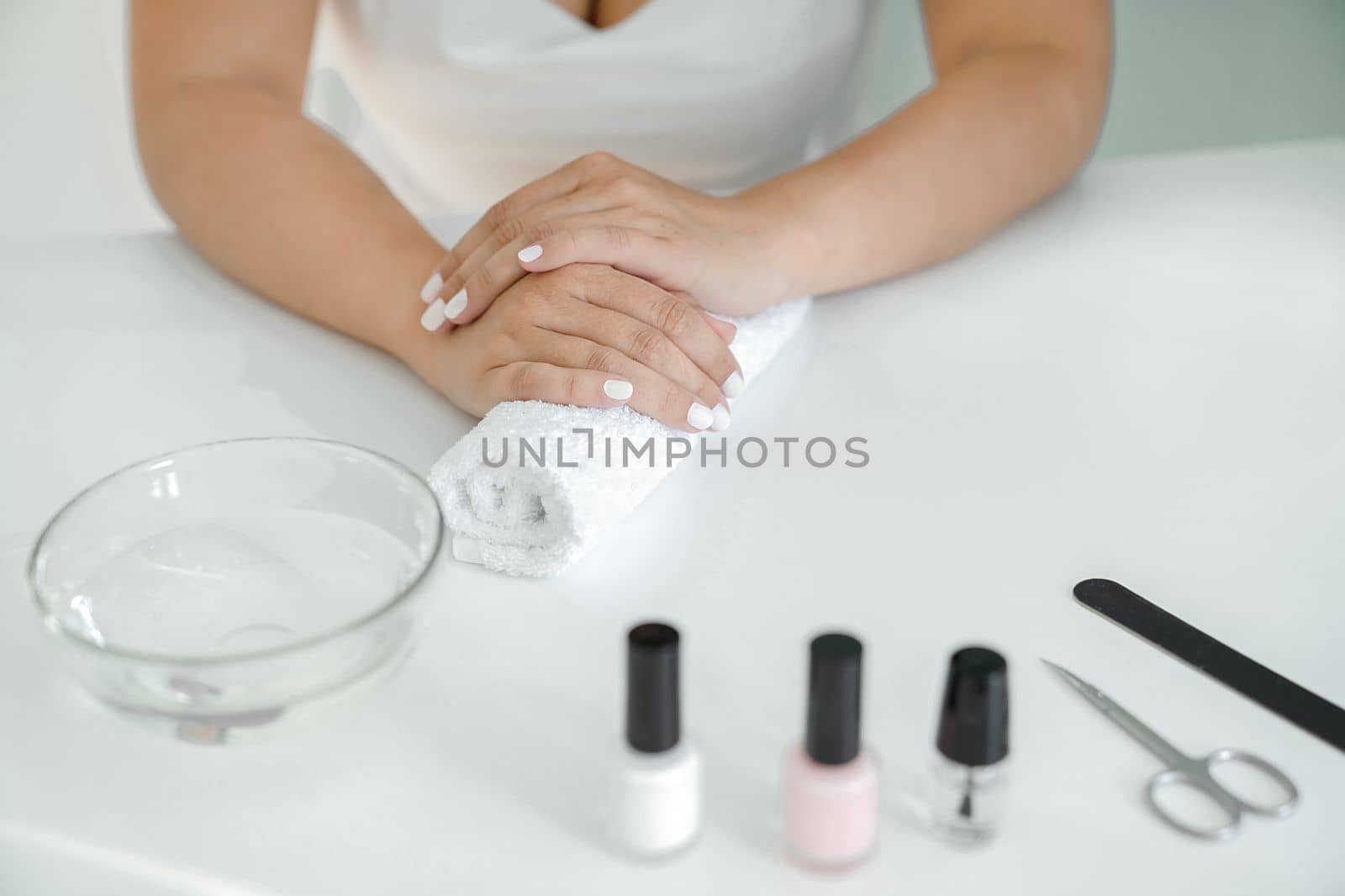 Women's hands with white manicure are lying on towel roller, waiting for varnish to dry, manicure accessories are lying on table next to it. Hands close-up, front view, without face