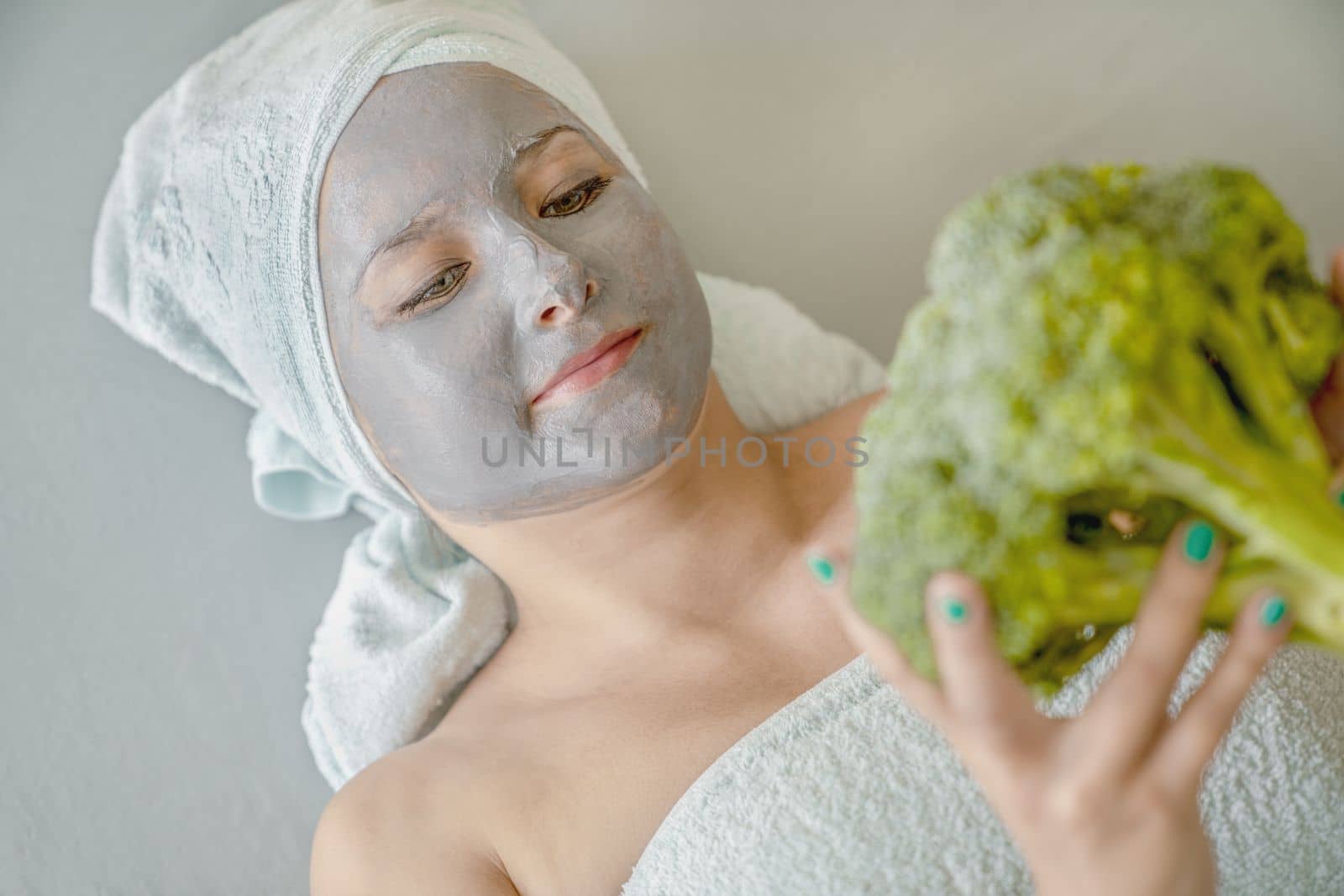 Funny young woman with cosmetic mask made of gray clay, her hair wrapped in towels, she holds broccoli in her hands, portrait, close-up. Spa procedure, concept of natural facial skin care. by Laguna781