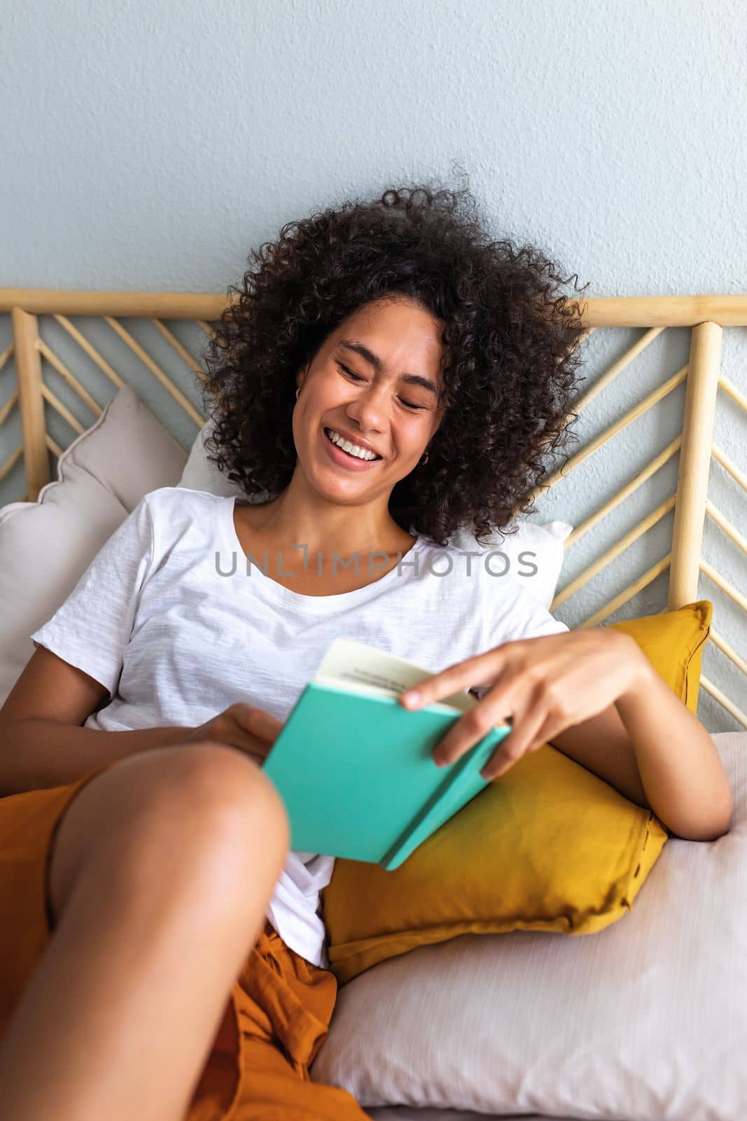 Vertical portrait of young African american woman with curly hair reading a book, laughing sitting comfortably on cozy bed. Lifestyle concept.