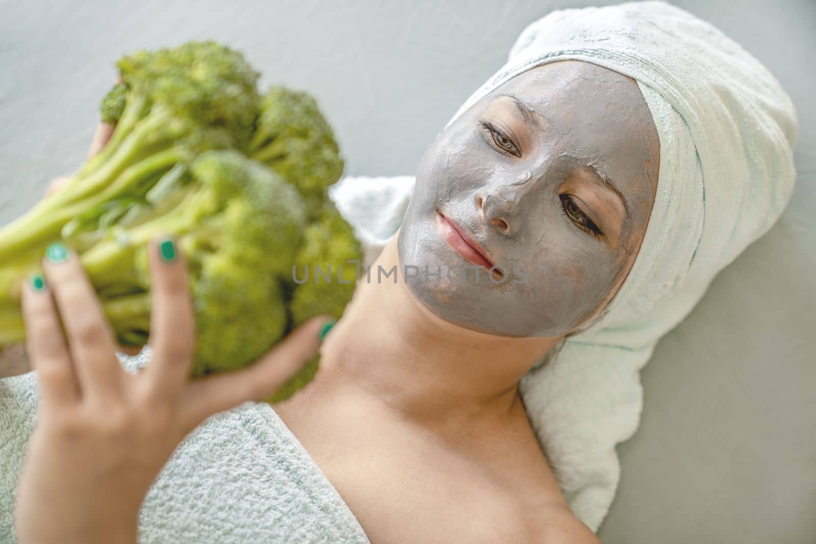 Concept of natural facial skin care. Funny young woman with cosmetic mask made of gray clay, her hair and body wrapped in towels, she holds broccoli in hands, portrait, close-up. Spa procedure. by Laguna781