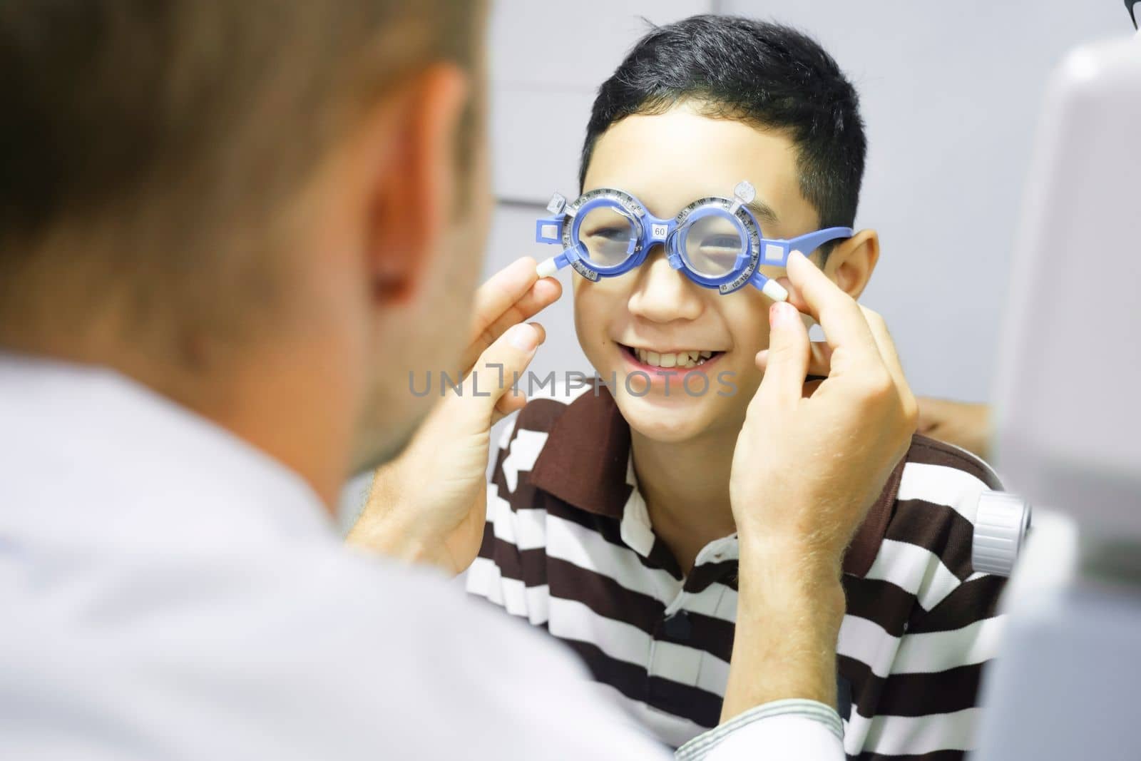 Smart young boy sitting in optometrist cabinet having his eyesight checking, examining, testing with trial frame glasses by professional optician for new pairs of eyeglasses.