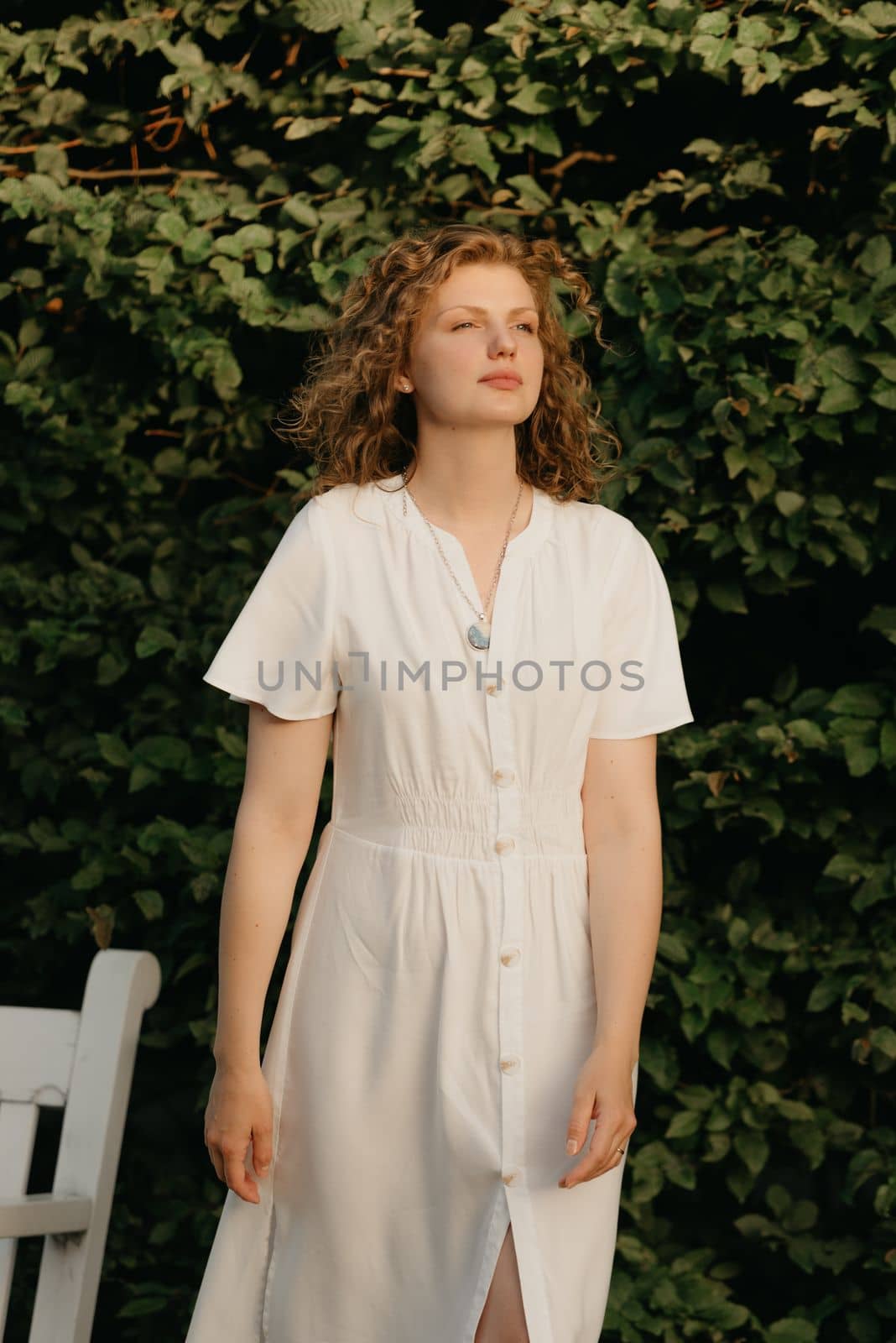 A woman with curly hair is posing in the garden in the evening. A lady in a white dress with green leaves in the background