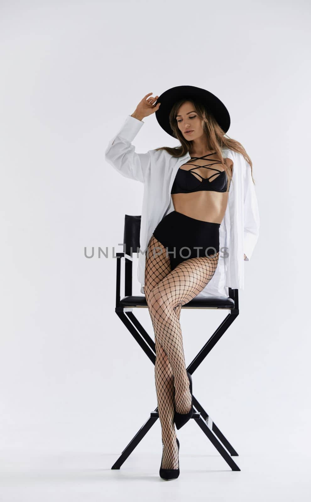 Beautiful brunette girl sits on chair and flirts, she is dressed in mesh tights, white shirt and black hat, long hair, pure white background, soft light. High quality photo