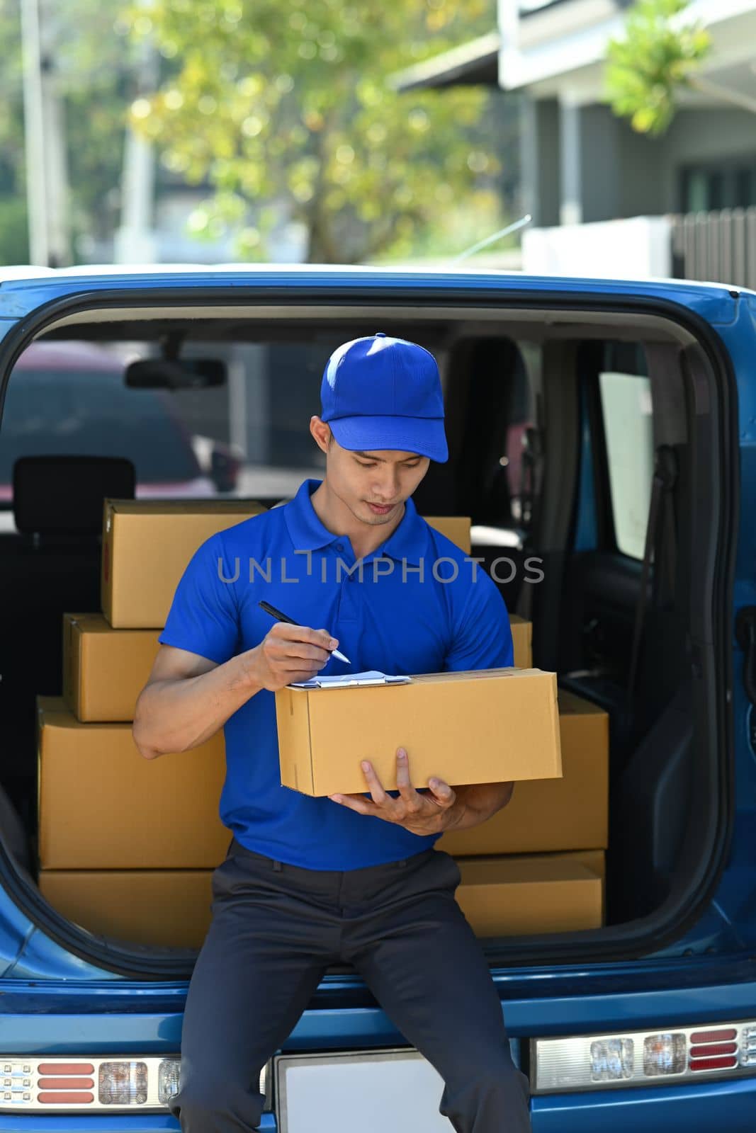 Delivery man in blue uniform using digital tablet while standing at open van full of delivering package.