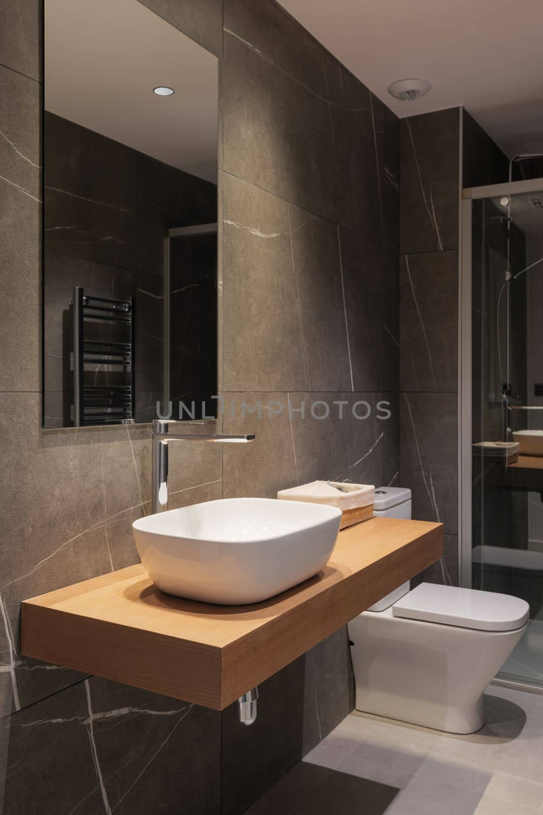 Beam from lamp illuminates white deep sink on marble countertop in noble wood color. Large mirror on wall reflects opposite side with heated towel rail. Modern design bathroom with dark marble walls. by apavlin