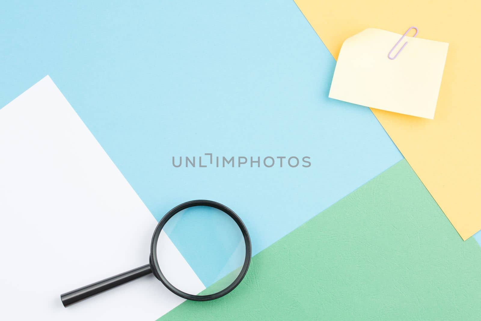 Colored paper background of geometric shapes with magnifying glass, note paper and paper clip. Office composition. Top view.