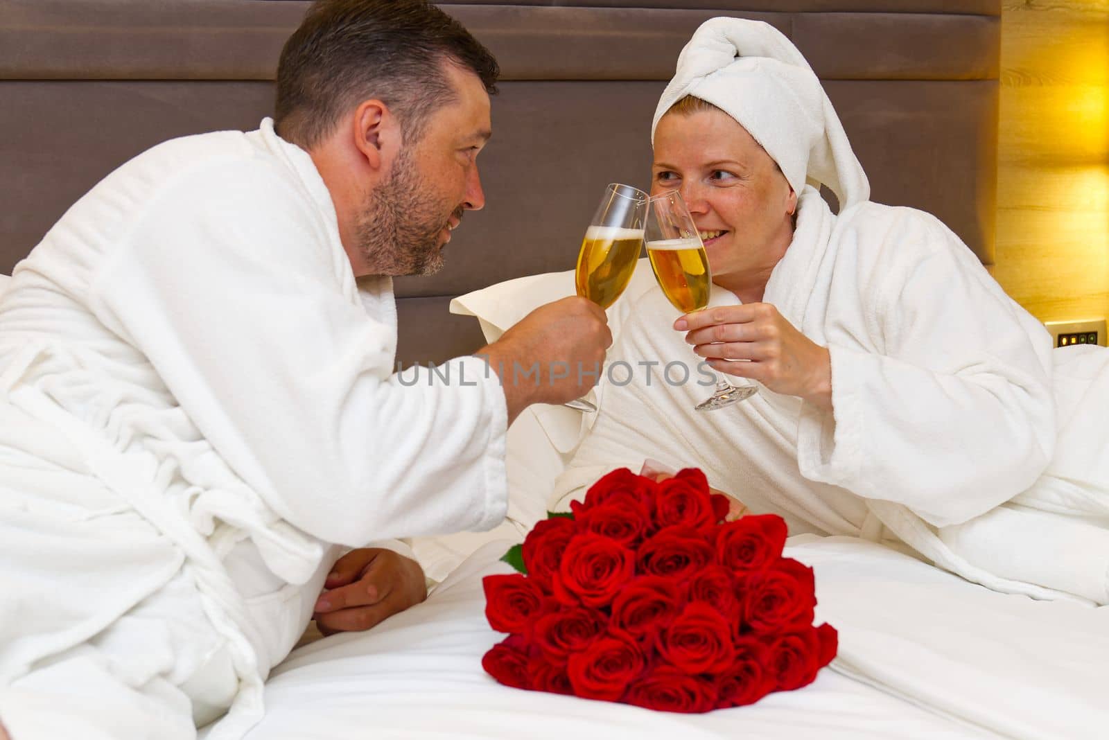 couple lies in bed and clink glasses with champagne, celebrate anniversary, celebrate birthday or romantic date of lovers. bouquet of red roses and lovers with glasses of champagne