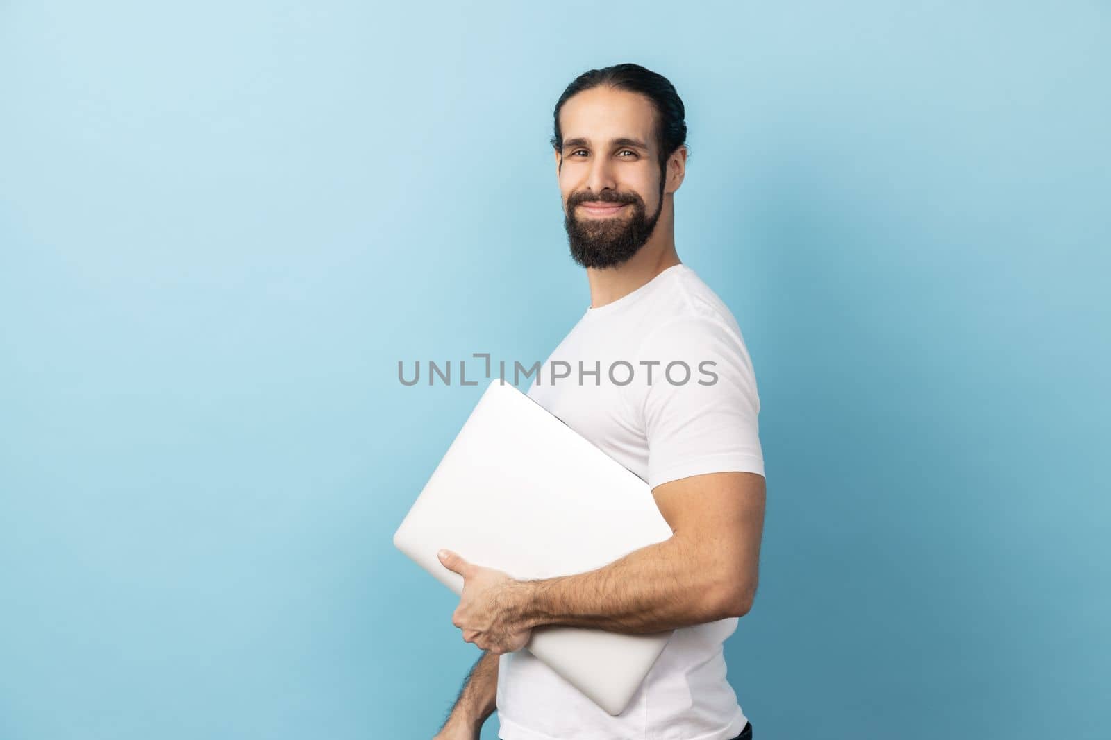Side view of man with beard wearing white T-shirt standing, holding closed laptop or folder, looking at camera with kind look, optimistic expression. Indoor studio shot isolated on blue background.