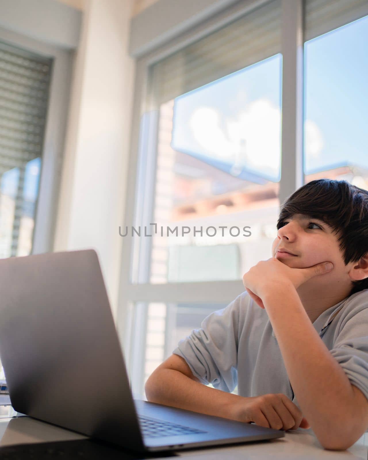 Kid sitting and thinking with laptop and table on window background at home.