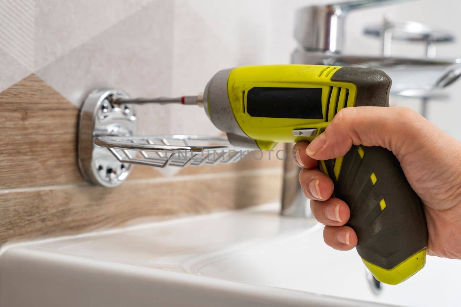 a close-up of an electric cordless screwdriver that wraps a screw into a hole in a ceramic tile to install a soap dish and shelves. home renovation. installing and fixing bathroom accessories