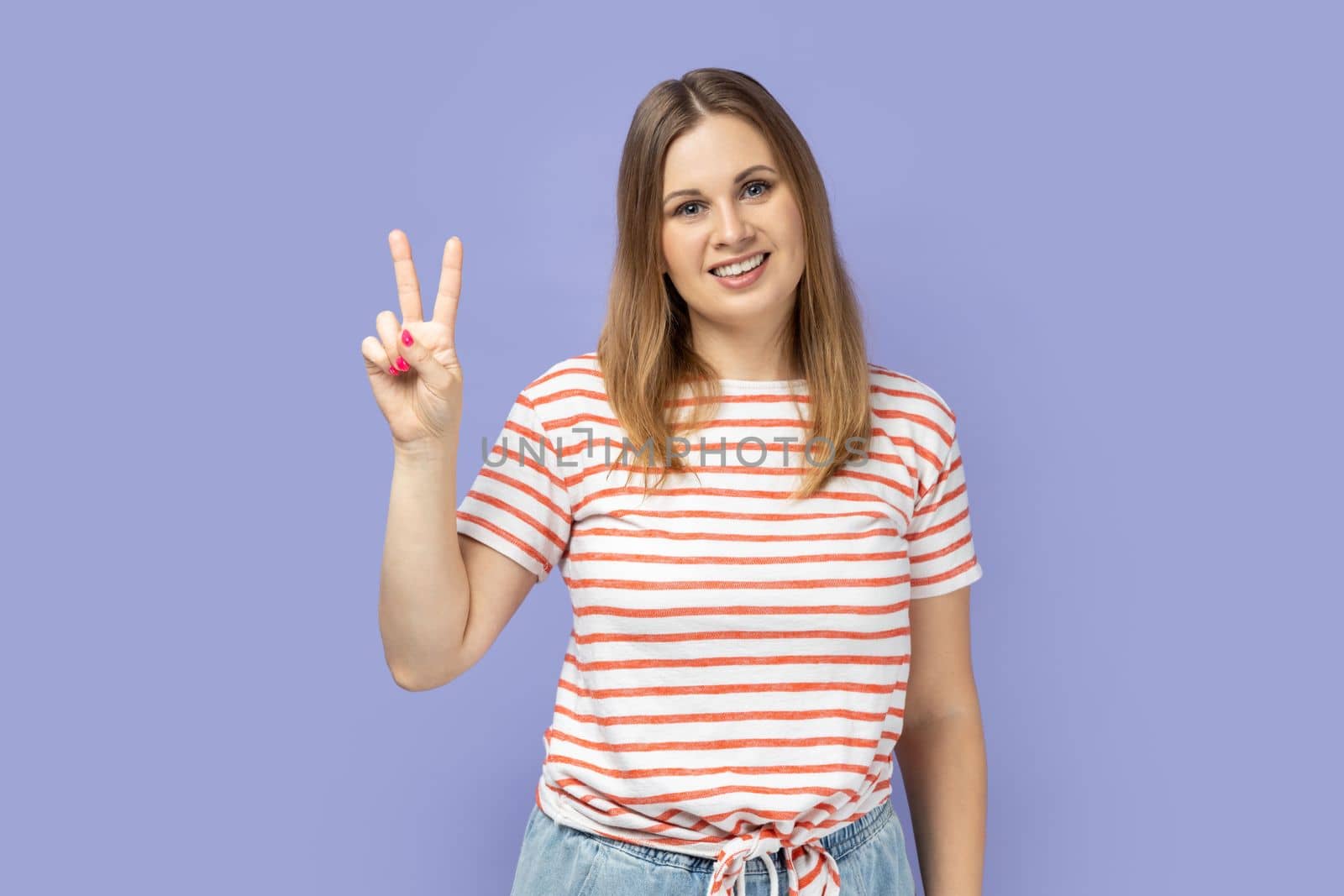 Portrait of beautiful blond woman wearing striped T-shirt smiling happily and showing v sign or peace gesture, rejoicing win. Indoor studio shot isolated on purple background.