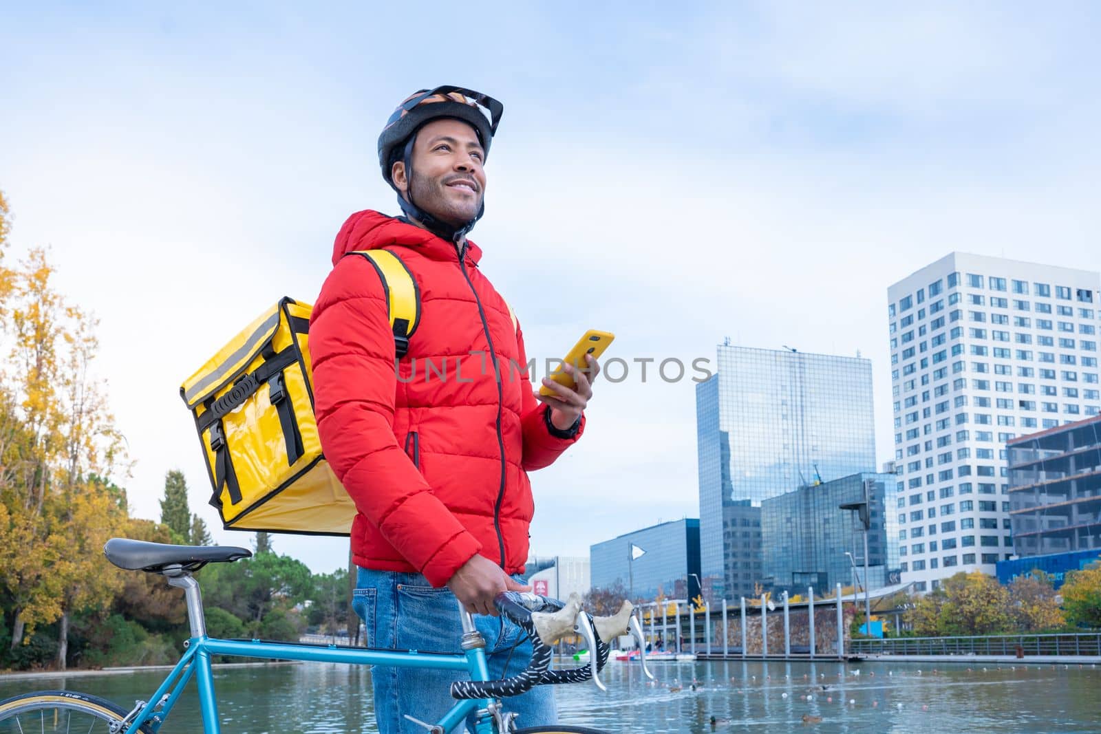 Food delivery concept. Young smiling African American driver with backpack looking smart phone in the city to deliver products for customers who order online purchases. Black male happy with his job.