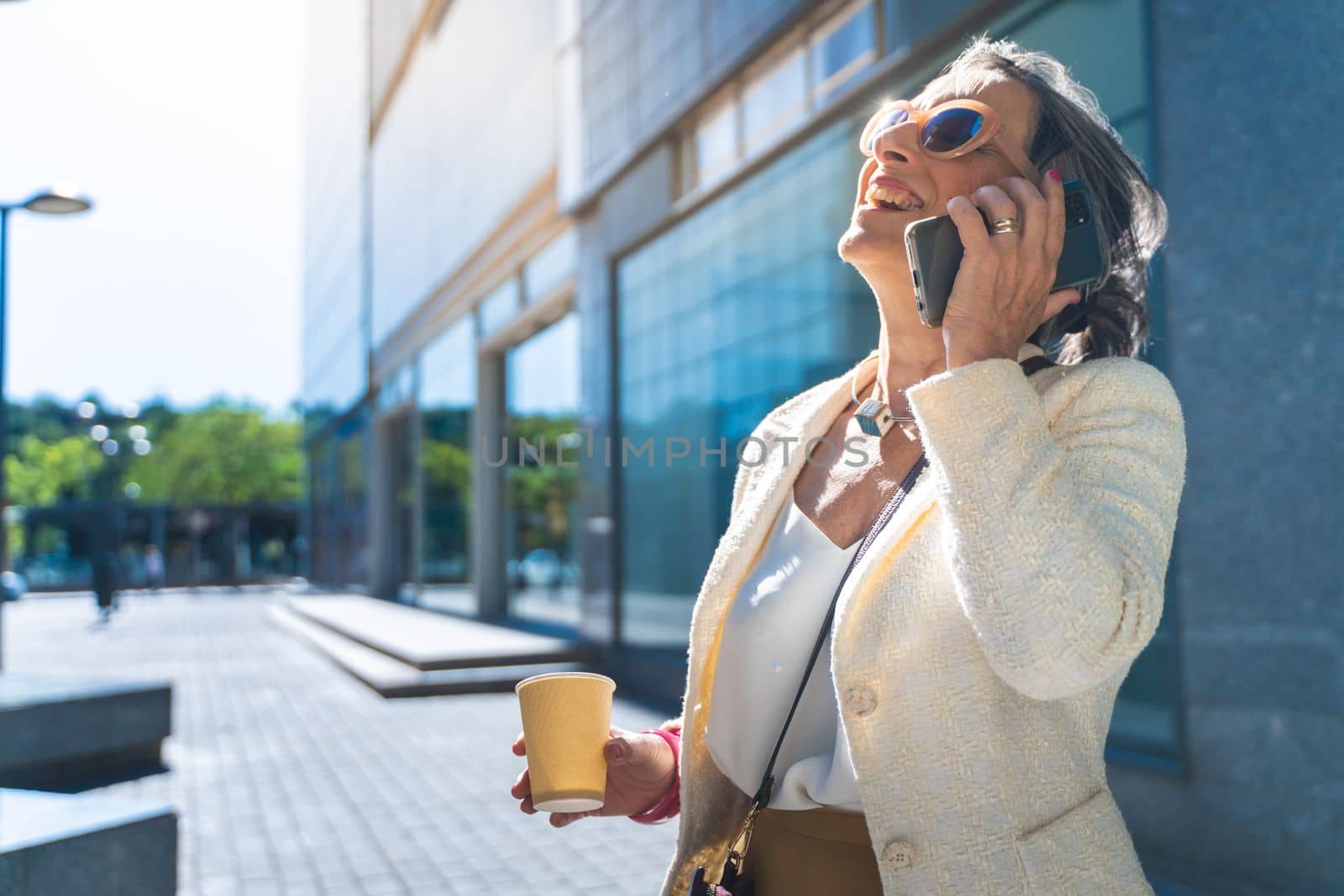 Attractive middle aged business woman with big smile talking with her smart phone with coffee cup in the street. by PaulCarr