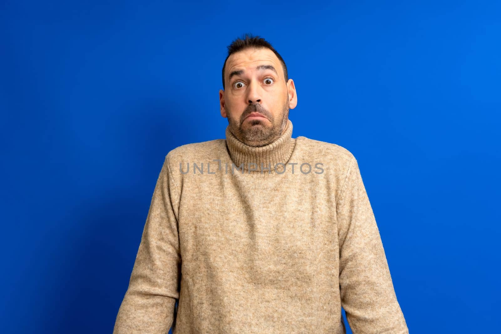 Frontal portrait of a hispanic man with a beard wearing a brown turtleneck doubting shrugging isolated on a blue background