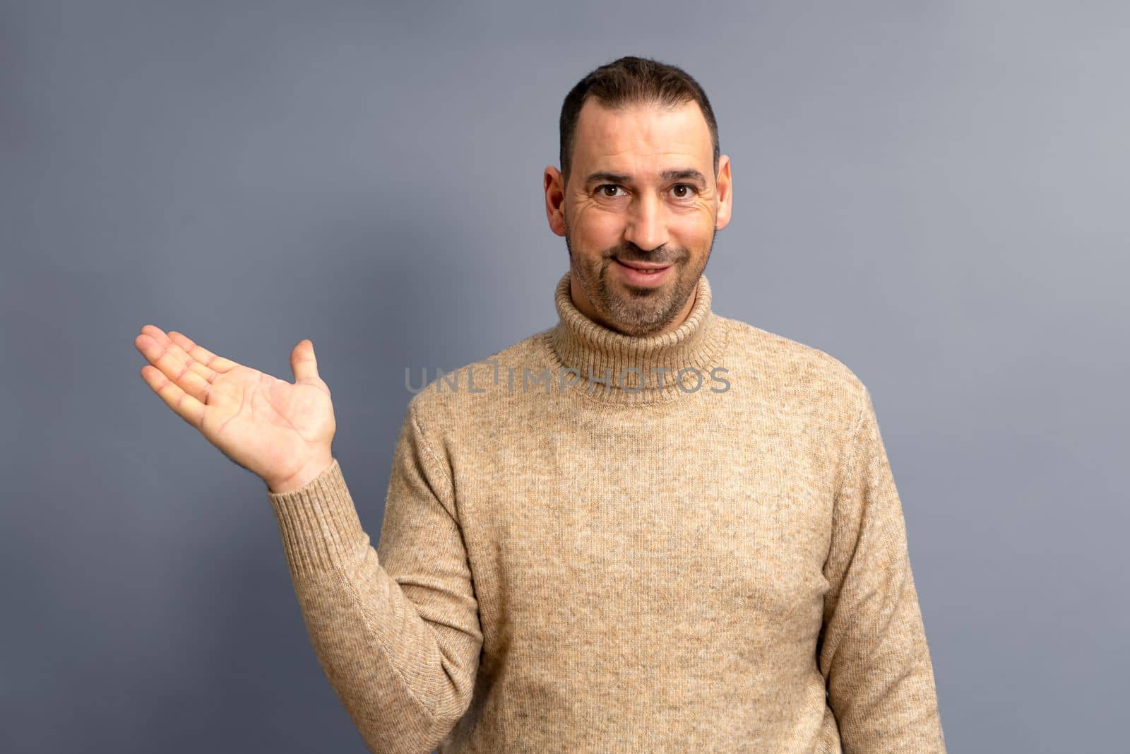 Hispanic man with beard and brown turtleneck sweater in friendly attitude offering something on the palm of his hand, isolated on gray background