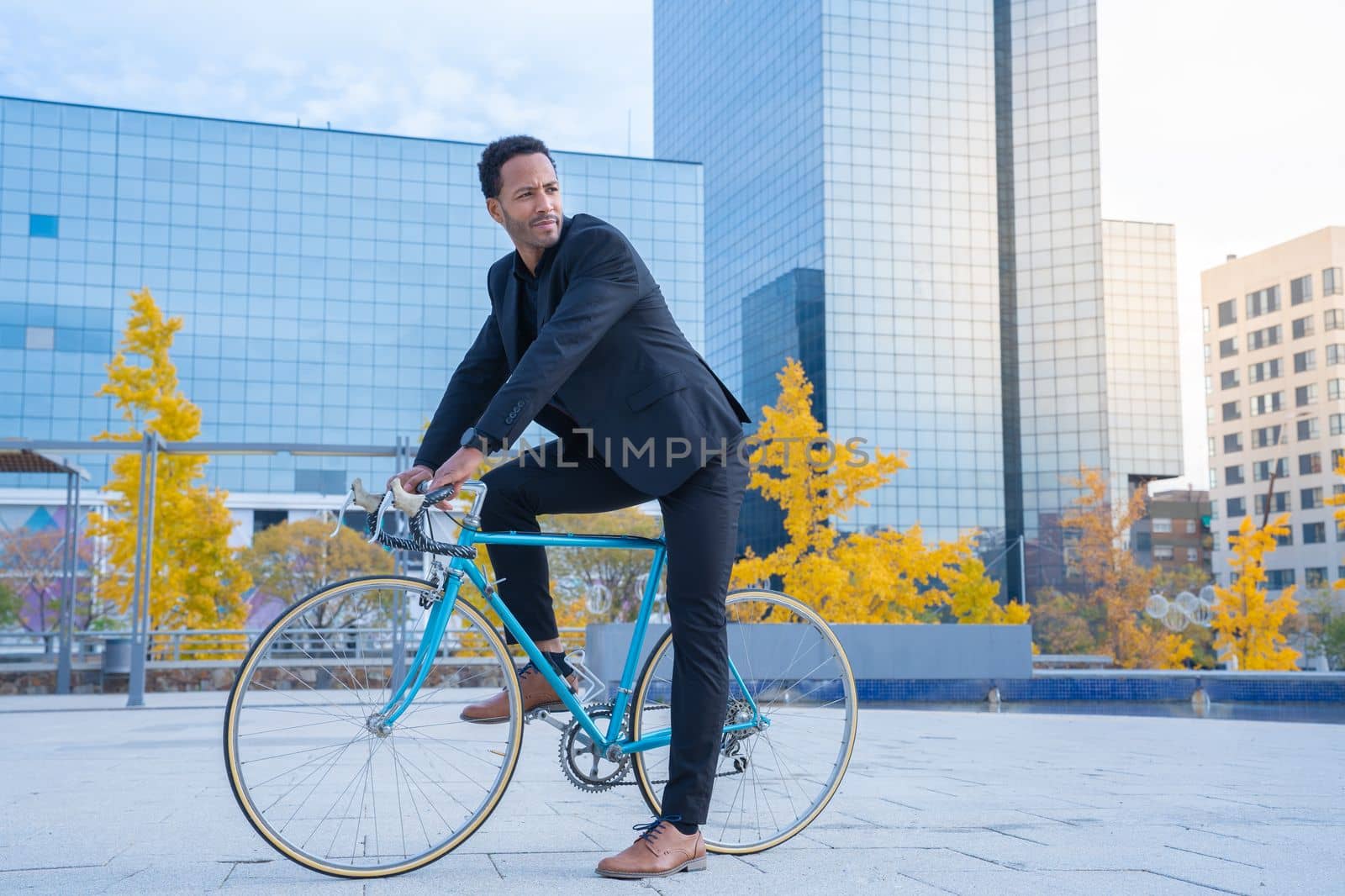 Young smiling stylish African American businessman in suit going to the office by bicycle in front of financial district. Business and transport concept. High quality vertical photo