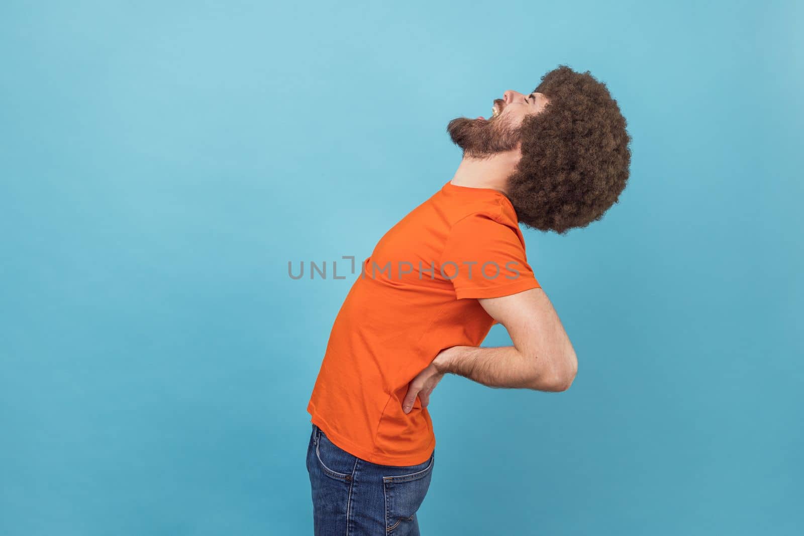 Side view portrait of man with Afro hairstyle wearing orange T-shirt standing with closed eyes, suffering lower back pain, kidney inflammation. Indoor studio shot isolated on blue background.