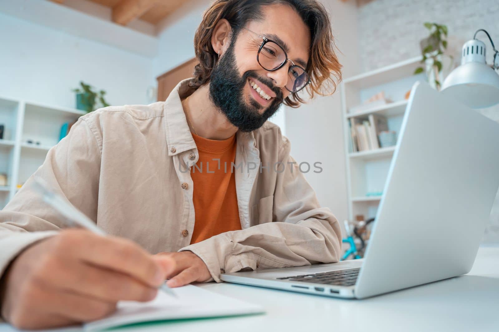 Happy young creative entrepreneur laughing smiling writes in a notebook next to laptop in a modern workspace office at home. Freelance guy starting small business. High quality photo