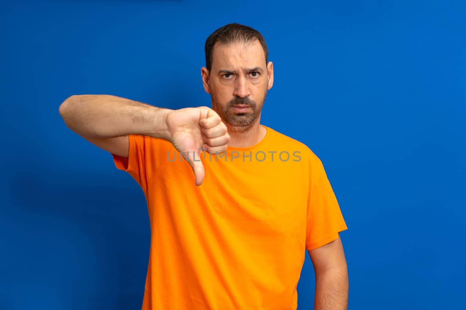 Handsome man wearing orange t-shirt standing over isolated blue background looking unhappy and angry showing rejection and negative gesture with thumb down. Bad expression.