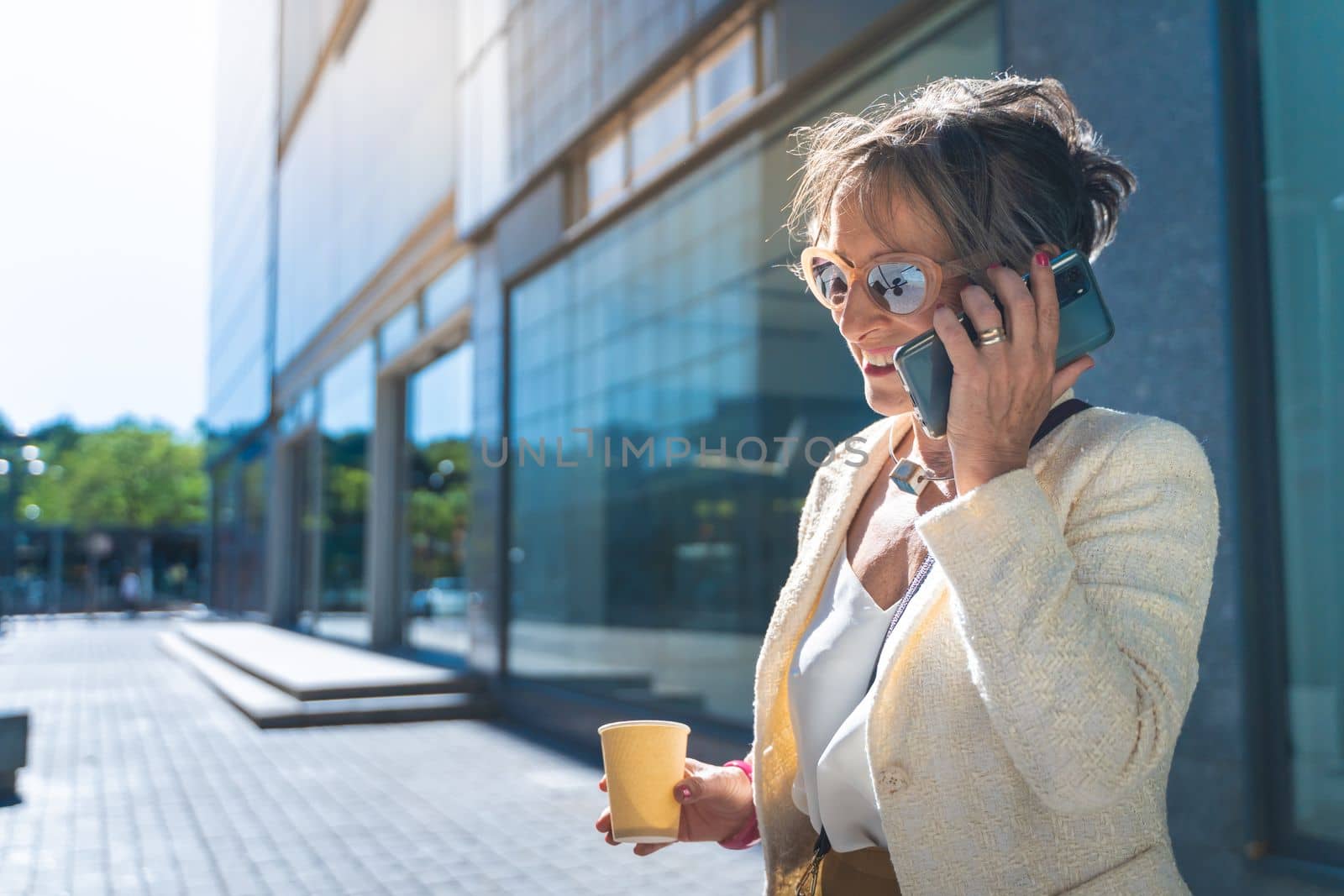 Attractive middle aged business woman smiling talking with her smart phone with coffee cup in the street. High quality photo