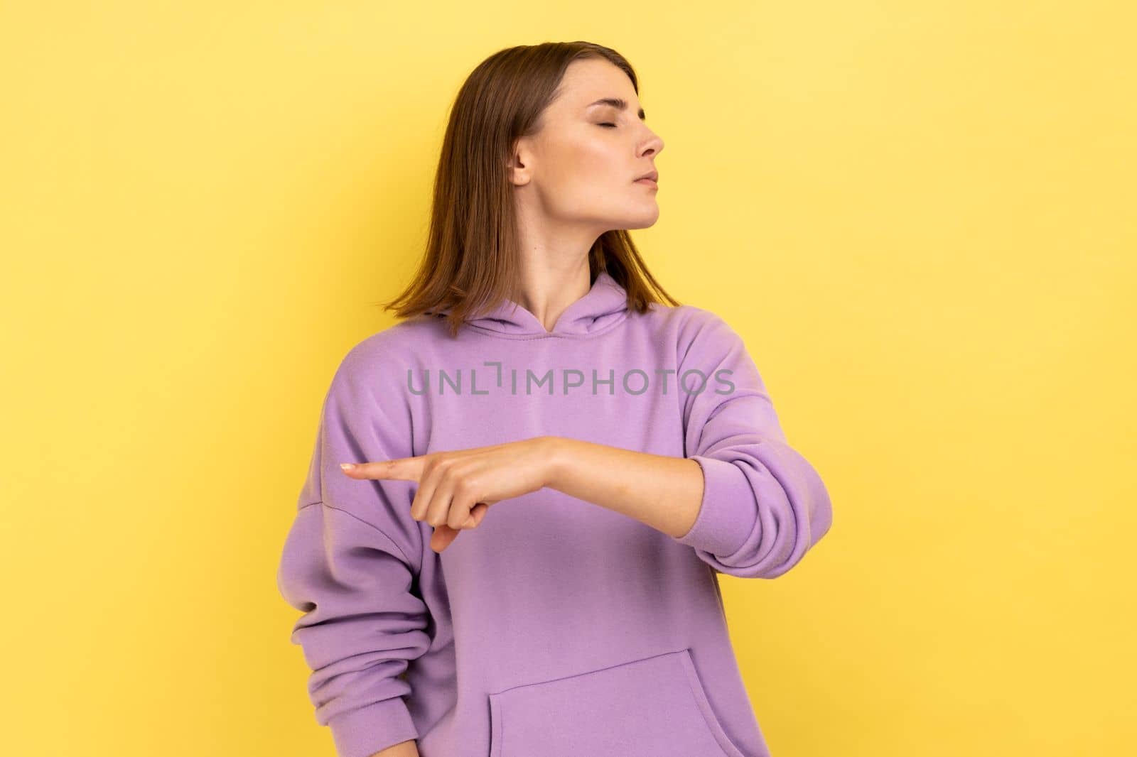 Get out. Portrait of upset vexed woman showing exit, demanding to leave her alone, has resentful irritated expression, wearing purple hoodie. Indoor studio shot isolated on yellow background.