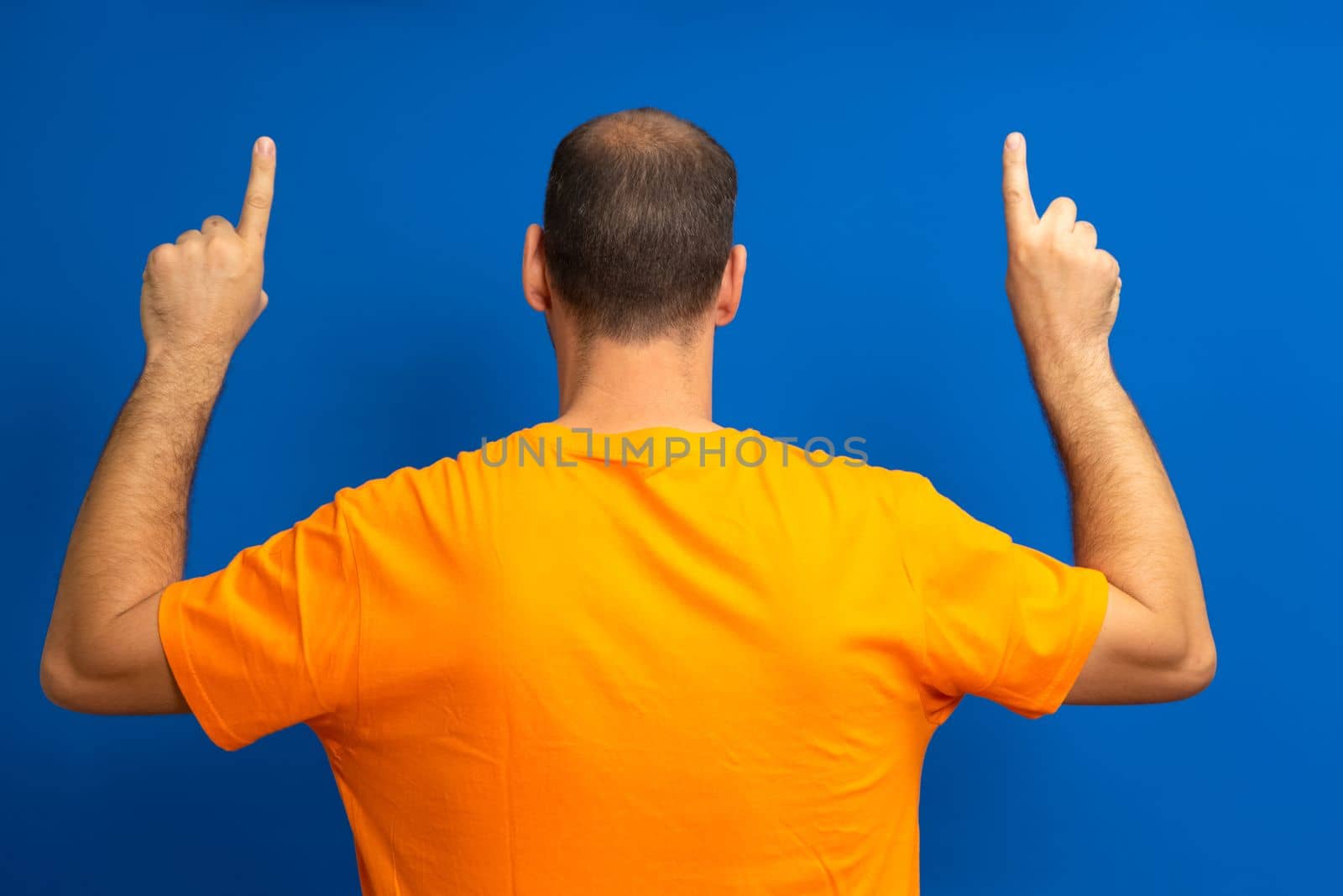Large man with receding hairline on his back pointing up with his index fingers isolated on blue studio background