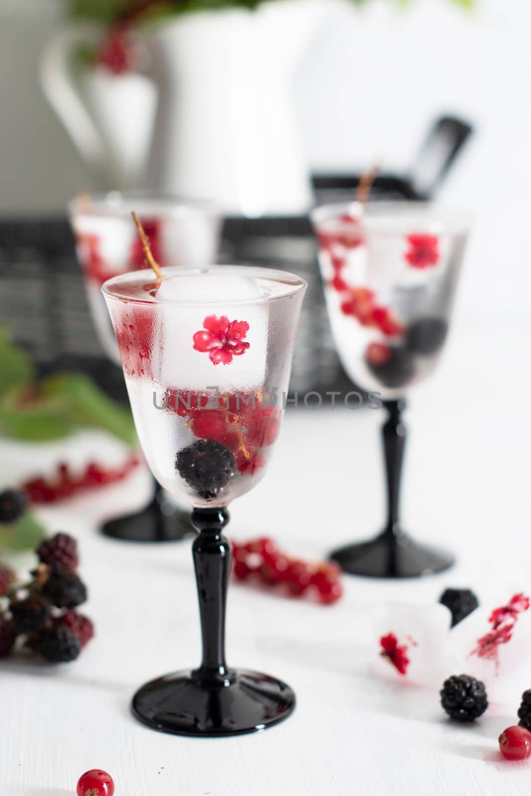 transparent gin and tonic with ice and fresh red currants and blackberries in beautiful crystal glasses against the background of a vase of flowers. High quality photo