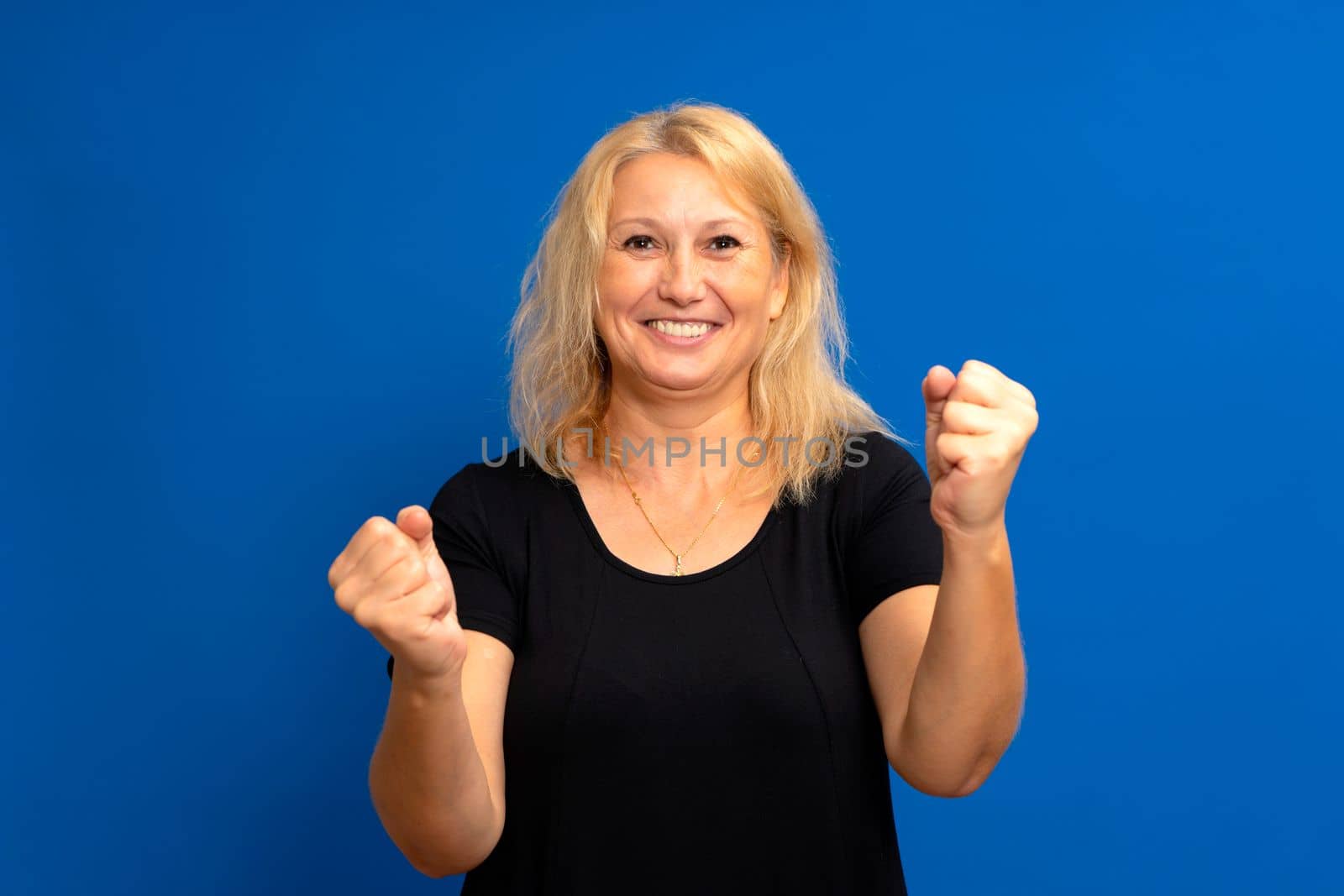Closeup of emotional attractive blonde caucasian woman in black t-shirt with hands in fists, isolated on blue background