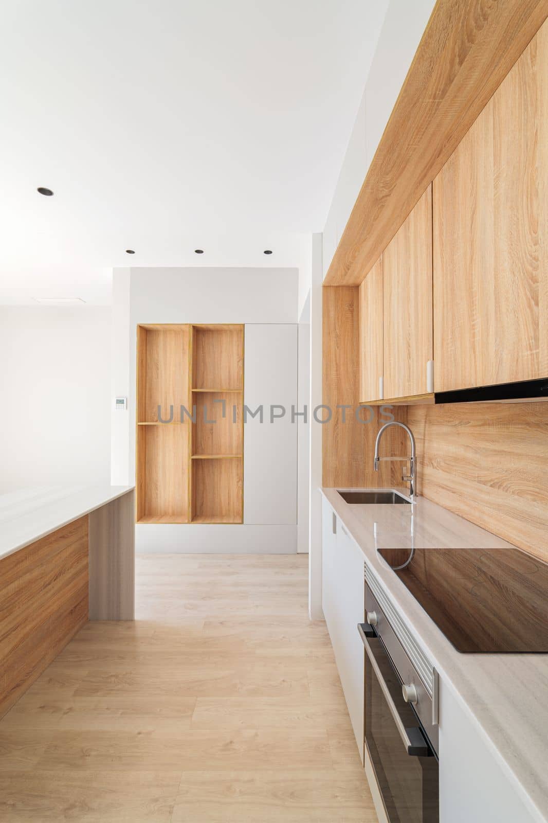 Along the wall is a modern kitchen with light wood cabinets. The marble top has an integrated induction hob and sink. In the doorway you can see a rack with shelves in the same style as the kitchen