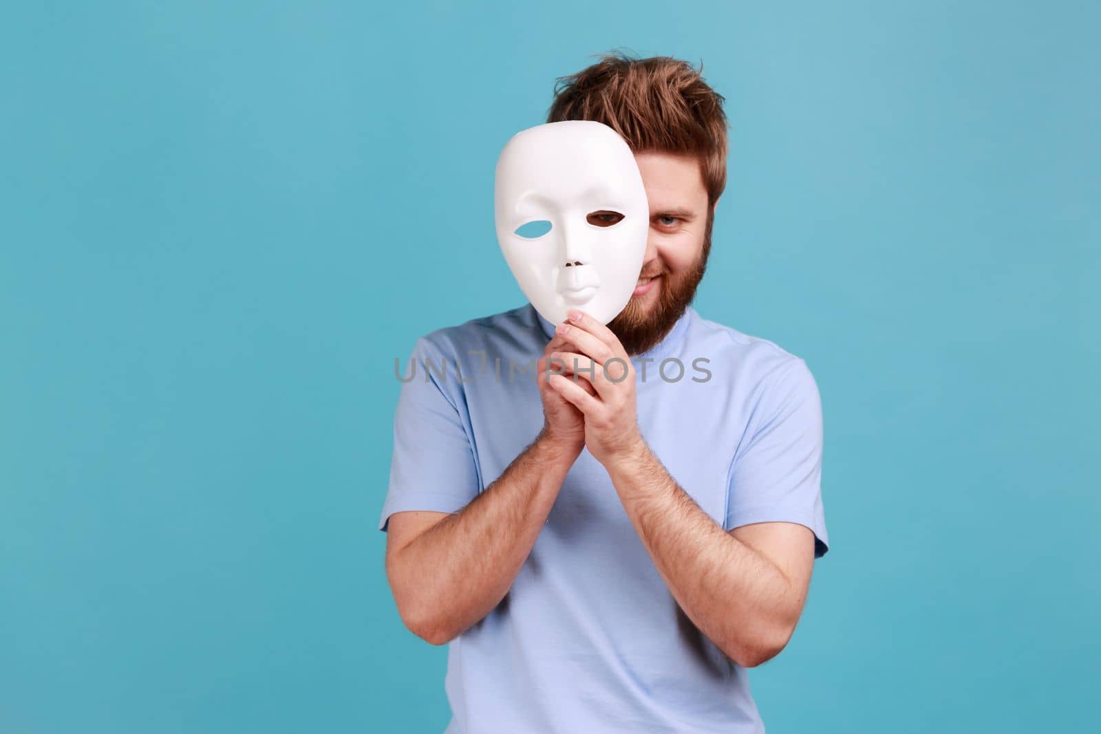 Portrait of positive bearded man removing white mask from face showing his smiling expression, good mood, pretending to be another person. Indoor studio shot isolated on blue background.