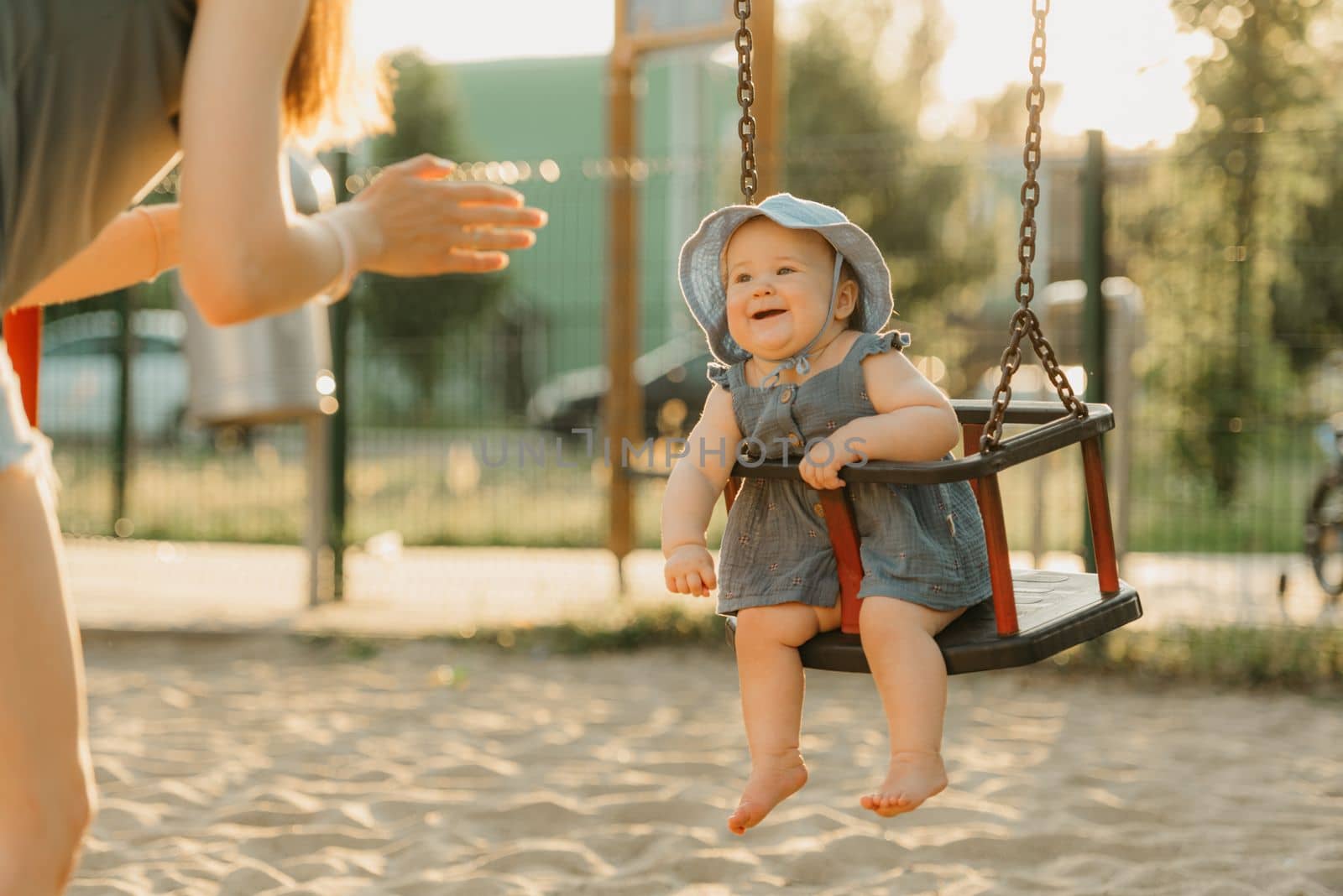 Toddler baby girl on a swing on the warm summer evening. Mother is swinging her young daughter on a sunny playground.