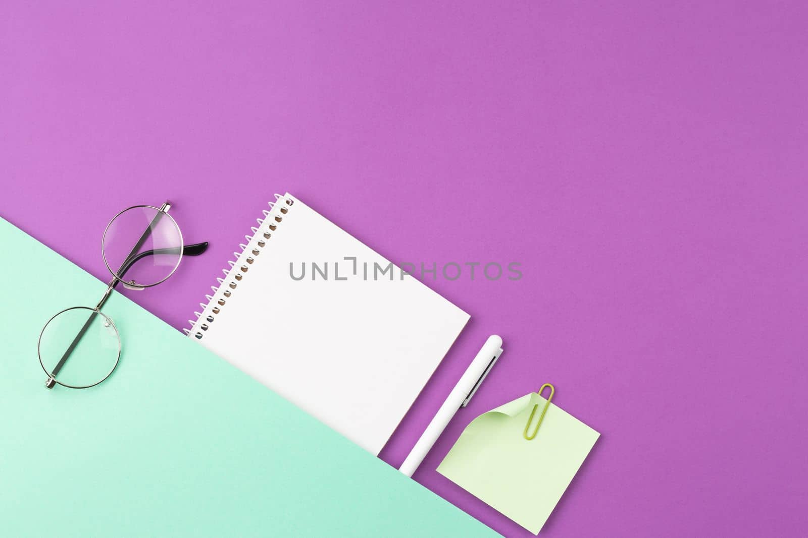 Open spiral notebook with white pen, glasses, note sheet and clip on two color background. Top view, flat lay.
