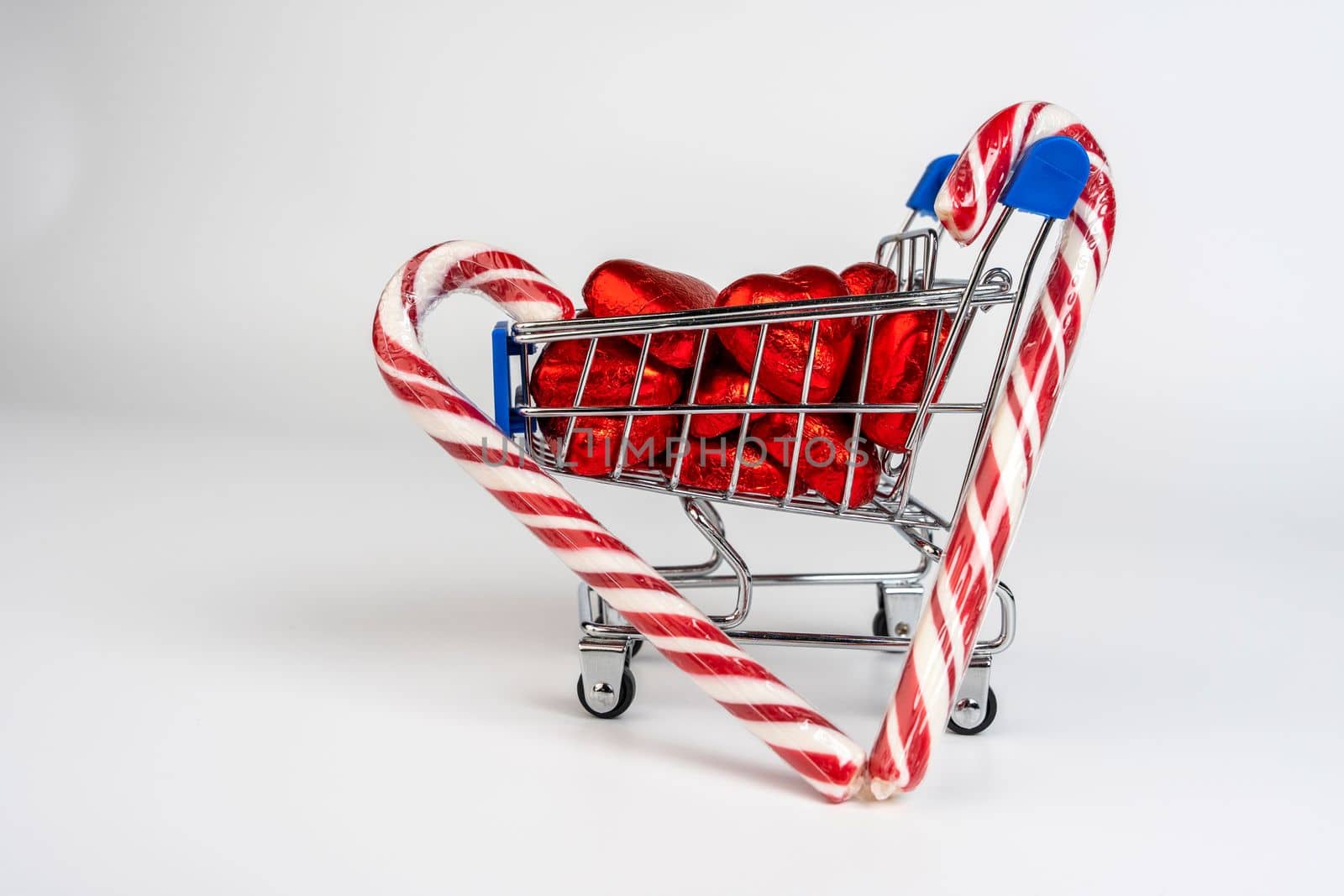 A supermarket trolley loaded with heart-shaped candies and Christmas caramel canes on a white background. Gifts and purchases for the New Year and Christmas. Online trading. A toy shopping cart carries a Christmas candy cane