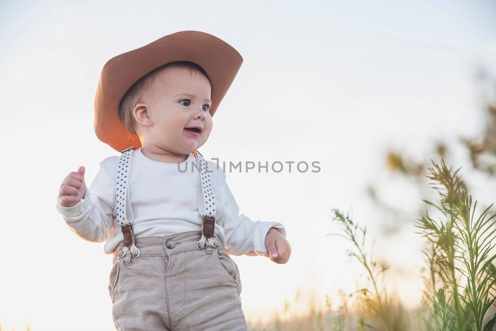 baby in a hat and suspenders looking in the field at sunset.