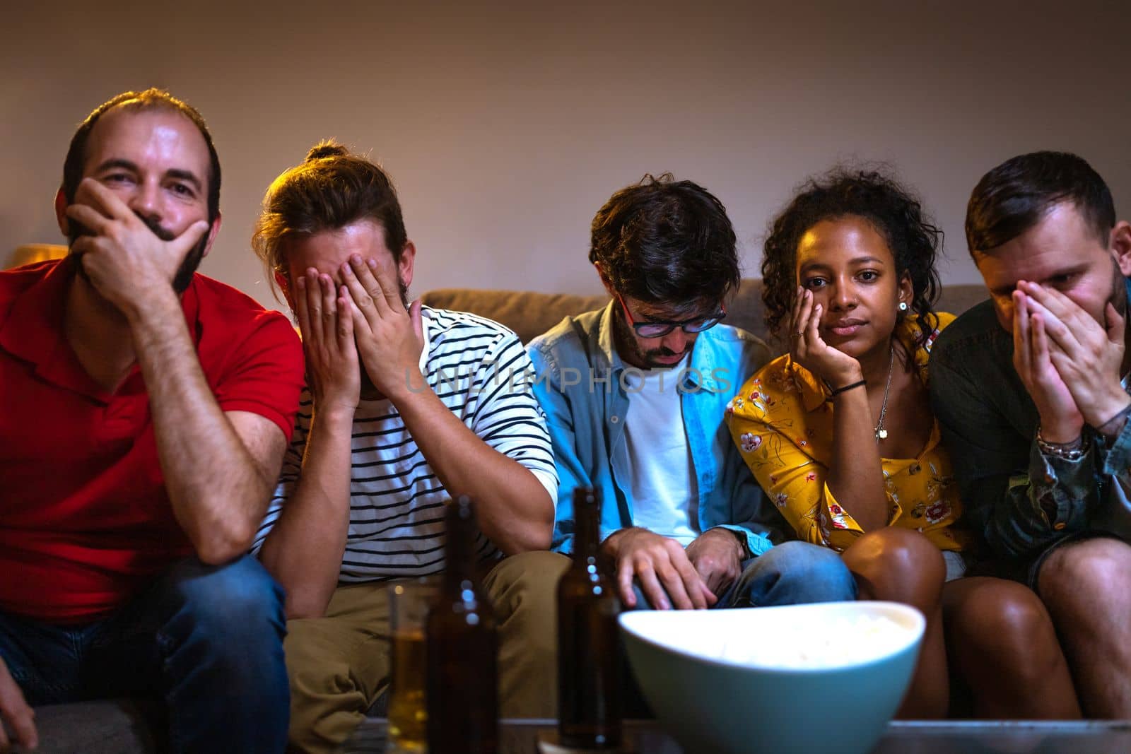 Group of friends watching football game on tv worried and sad because their team is losing. Soccer. Looking at camera. by Hoverstock