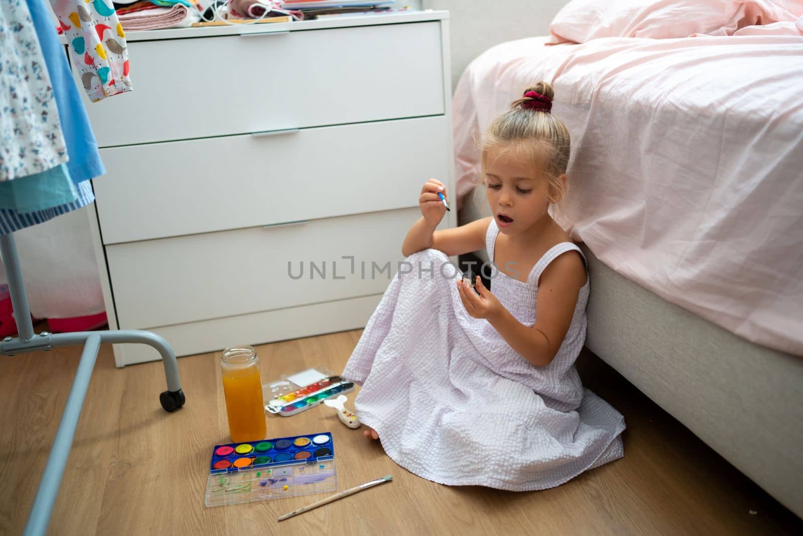 Caucasian child girl is painting watercolor paint on small ceramic figure with fun. Kids arts and crafts creative activity class at home. Little girl make ceramic decor sitting floor