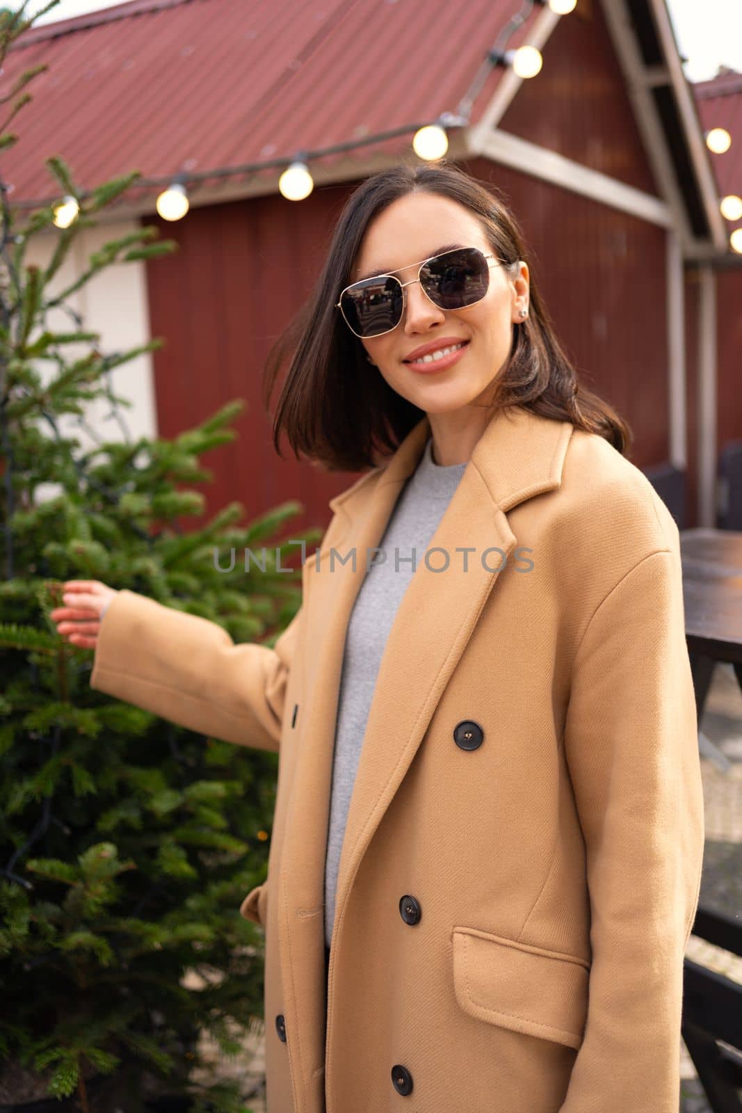 Woman in sunglasses dressed cream color coat standing outdoor with Christmas decoration on background choose Christmas tree. Happy positive woman walking city Christmas market vertical